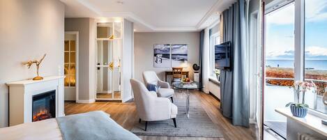 A modern hotel room with a fireplace, a comfortable sofa, a dining area, and a large window with a view of the sea.