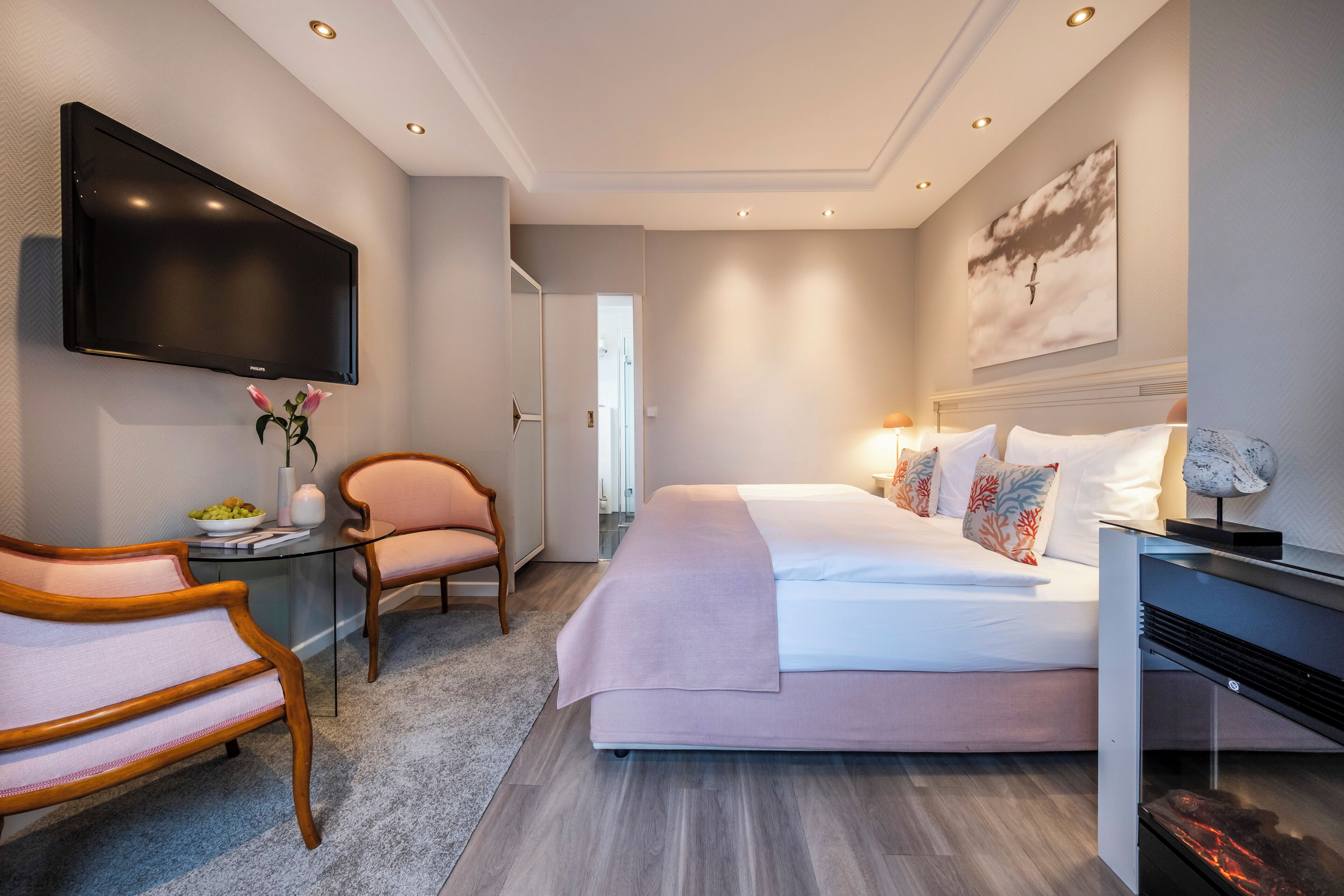 A modern hotel room with a large bed, a flat-screen TV mounted on the wall, a glass coffee table, and a seating area with a chair and a small table.
