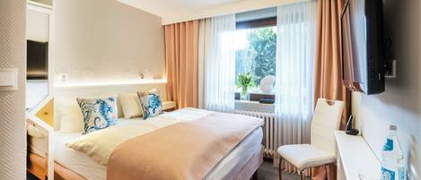 Una habitación de hotel con cama, escritorio, silla, televisor y ventana con cortinas.