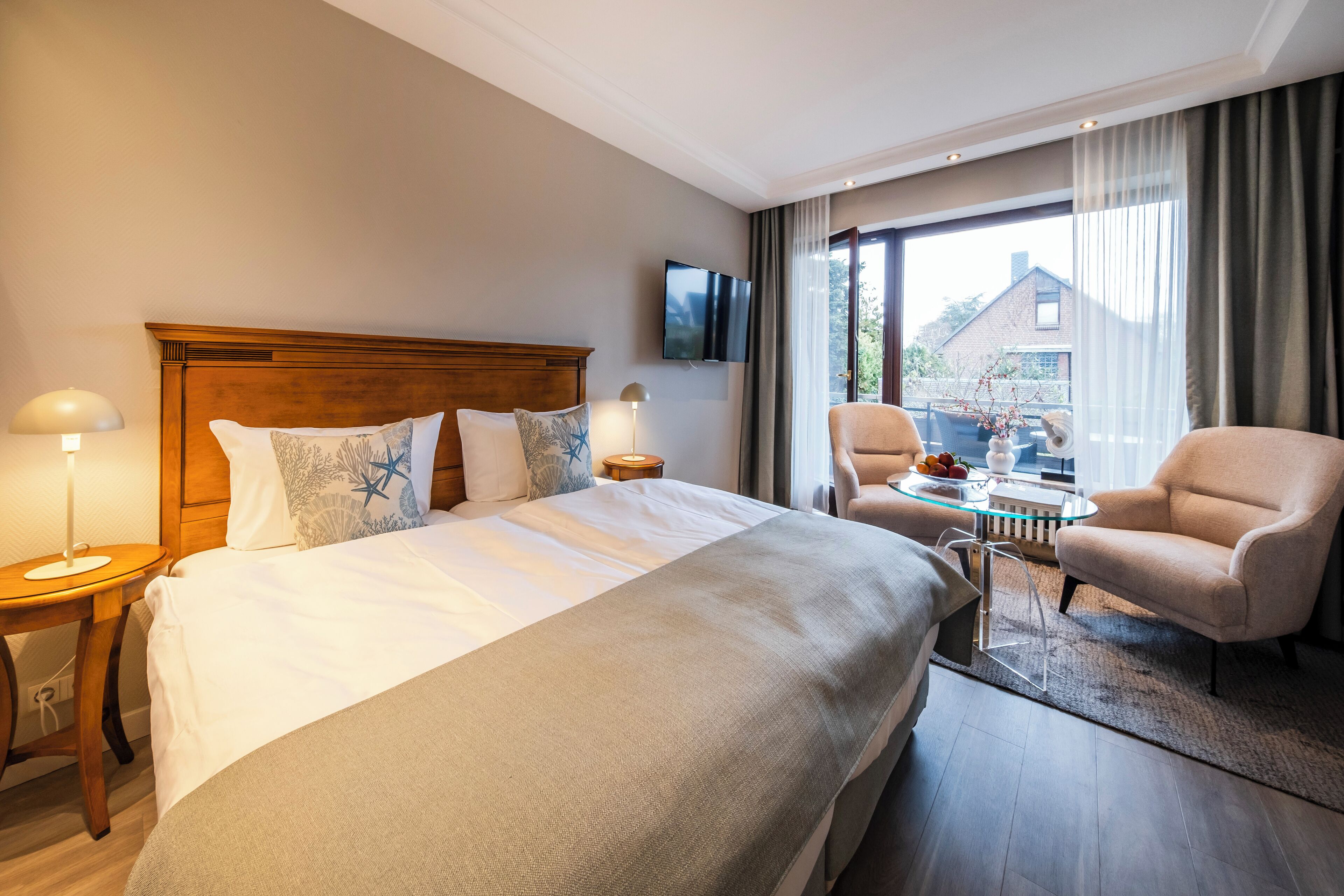 Habitación de hotel con una cama grande, televisor montado en la pared, zona de estar con mesa y sillas, y vistas al exterior.