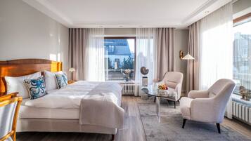 A modern hotel room with a large bed, a sitting area with a glass coffee table, and a view of the city through large windows.