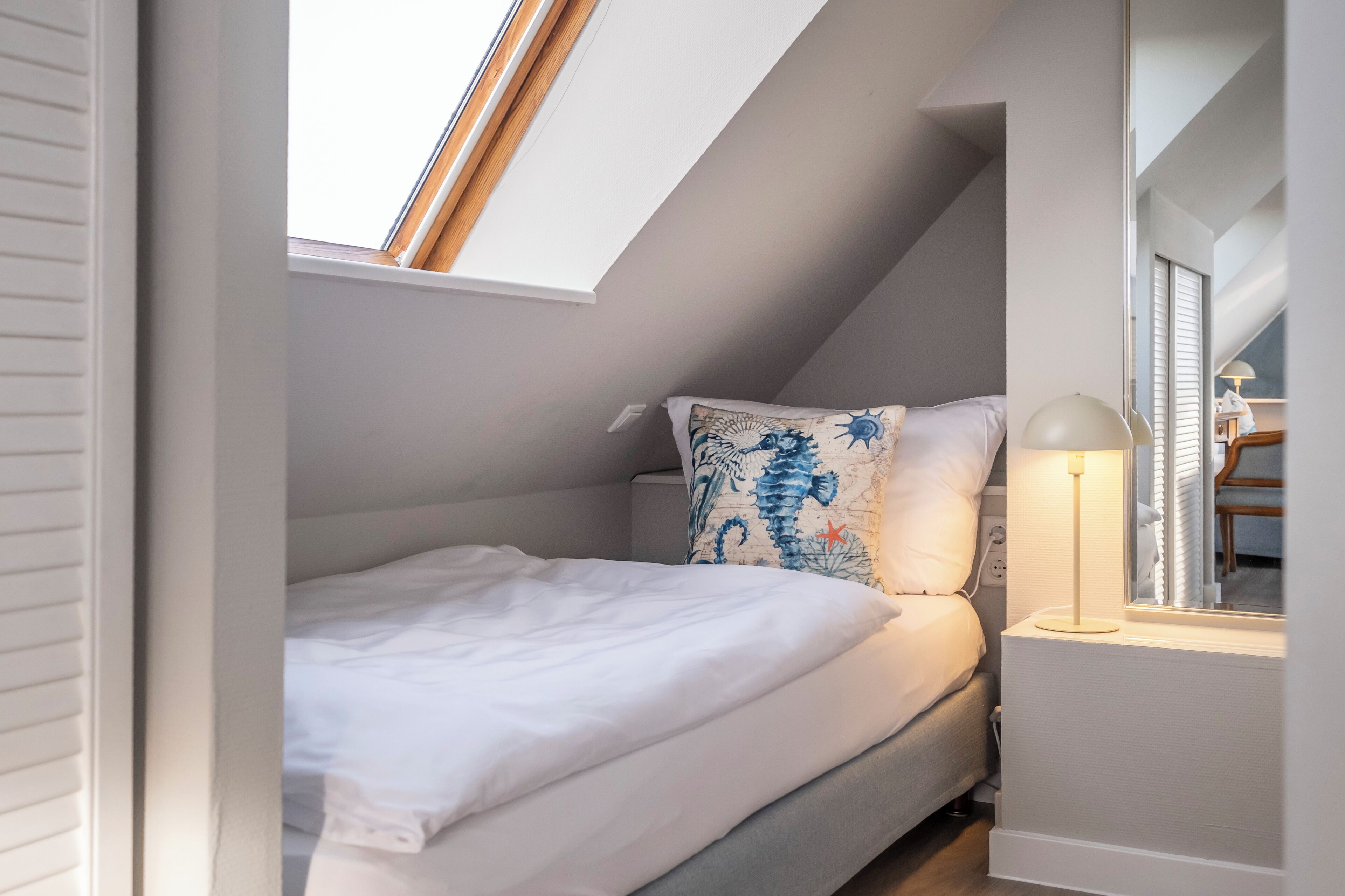 Een kleine, gezellige slaapkamer met dakraam, een bed met wit beddengoed, een decoratief kussen en een bedlampje.
