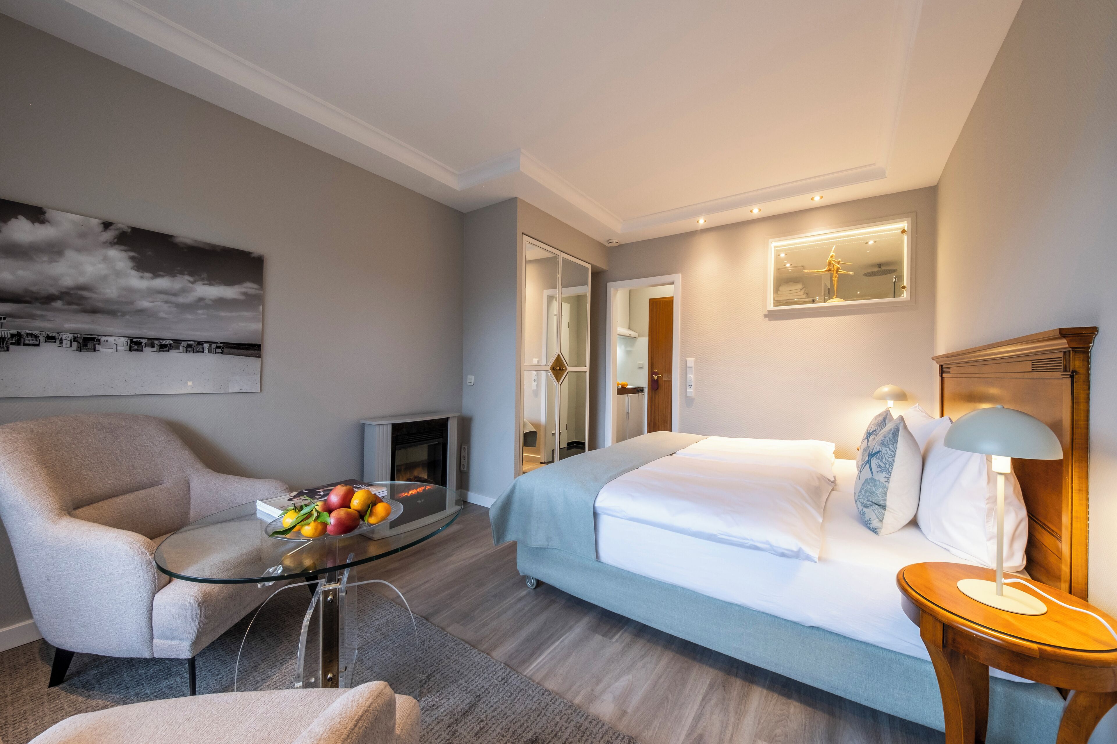 Une chambre d’hôtel avec un lit, une table en verre sur laquelle se trouvent des fruits, une chaise, une cheminée et une grande fenêtre.