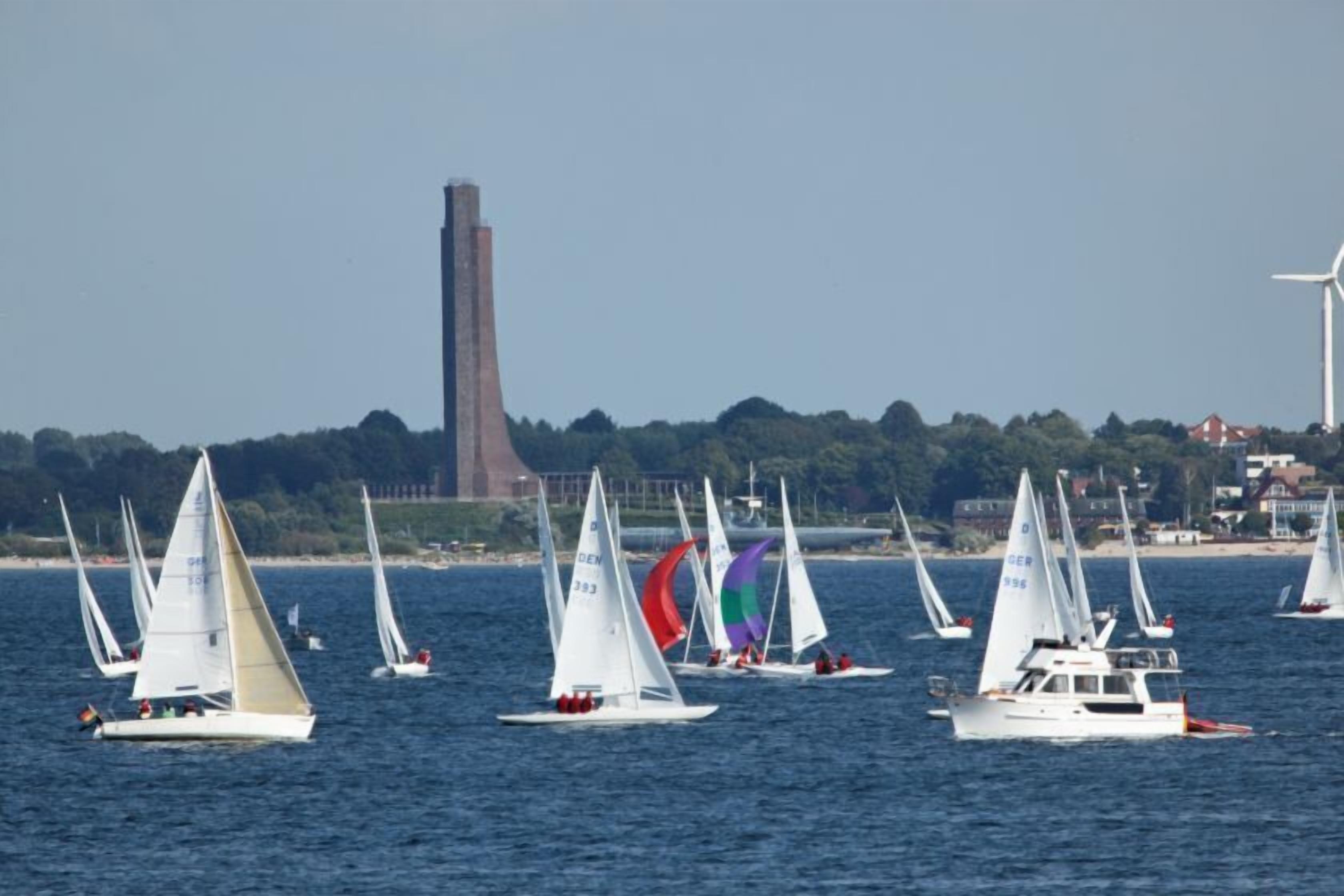Sailboats on a body of water with a distant view of a tall structure and wind turbines.