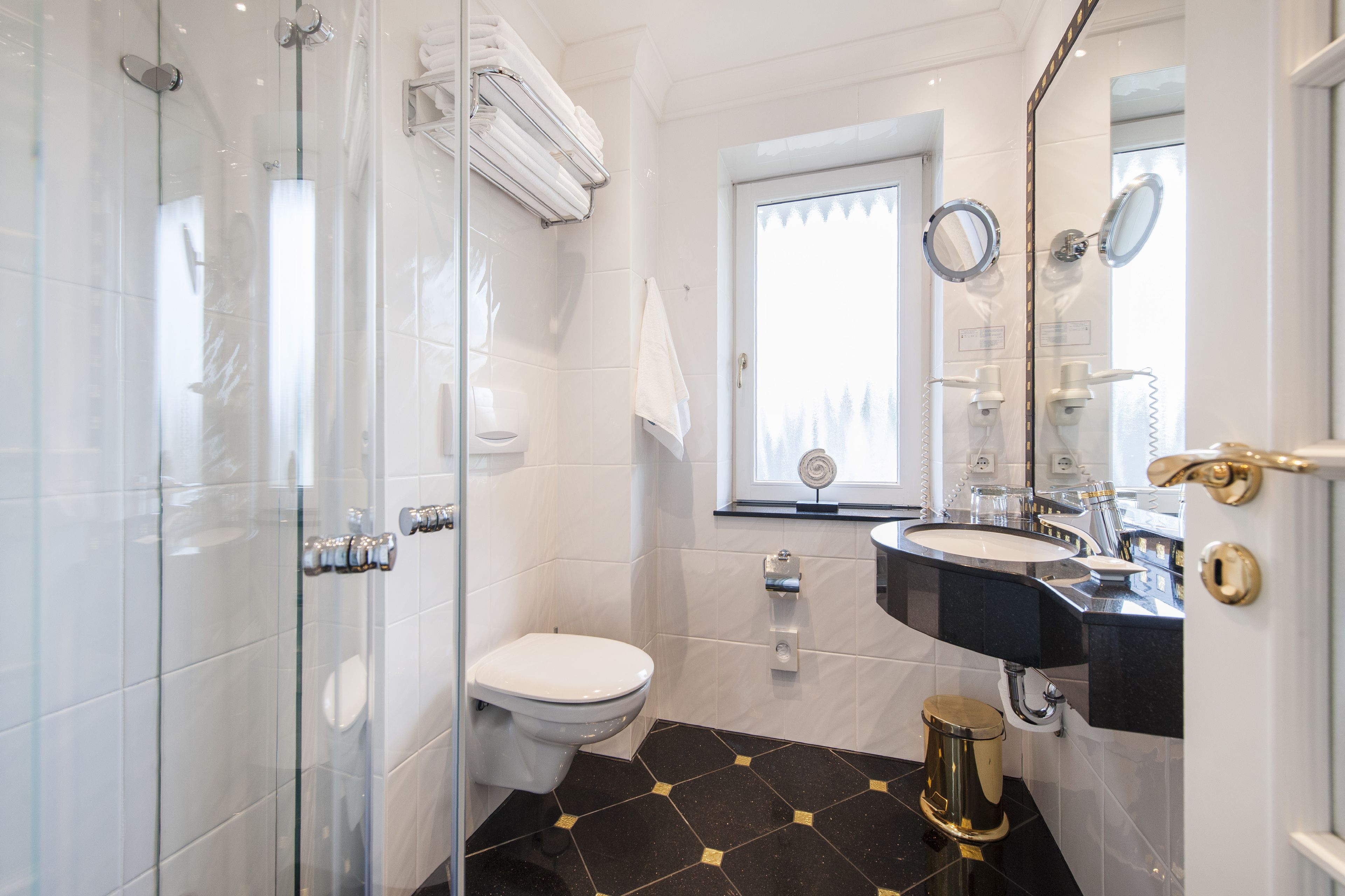 Een badkamer met een douchecabine van glas, een wit toilet, een wastafel met een zwart aanrechtblad en een spiegel met een gouden handgreep.