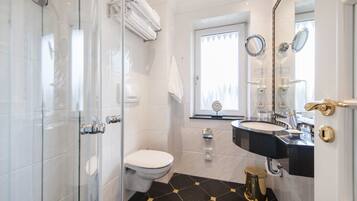 Et badeværelse med glasbrusekabine, hvidt toilet, vask med sort bordplade og spejl med forgyldt håndtag.