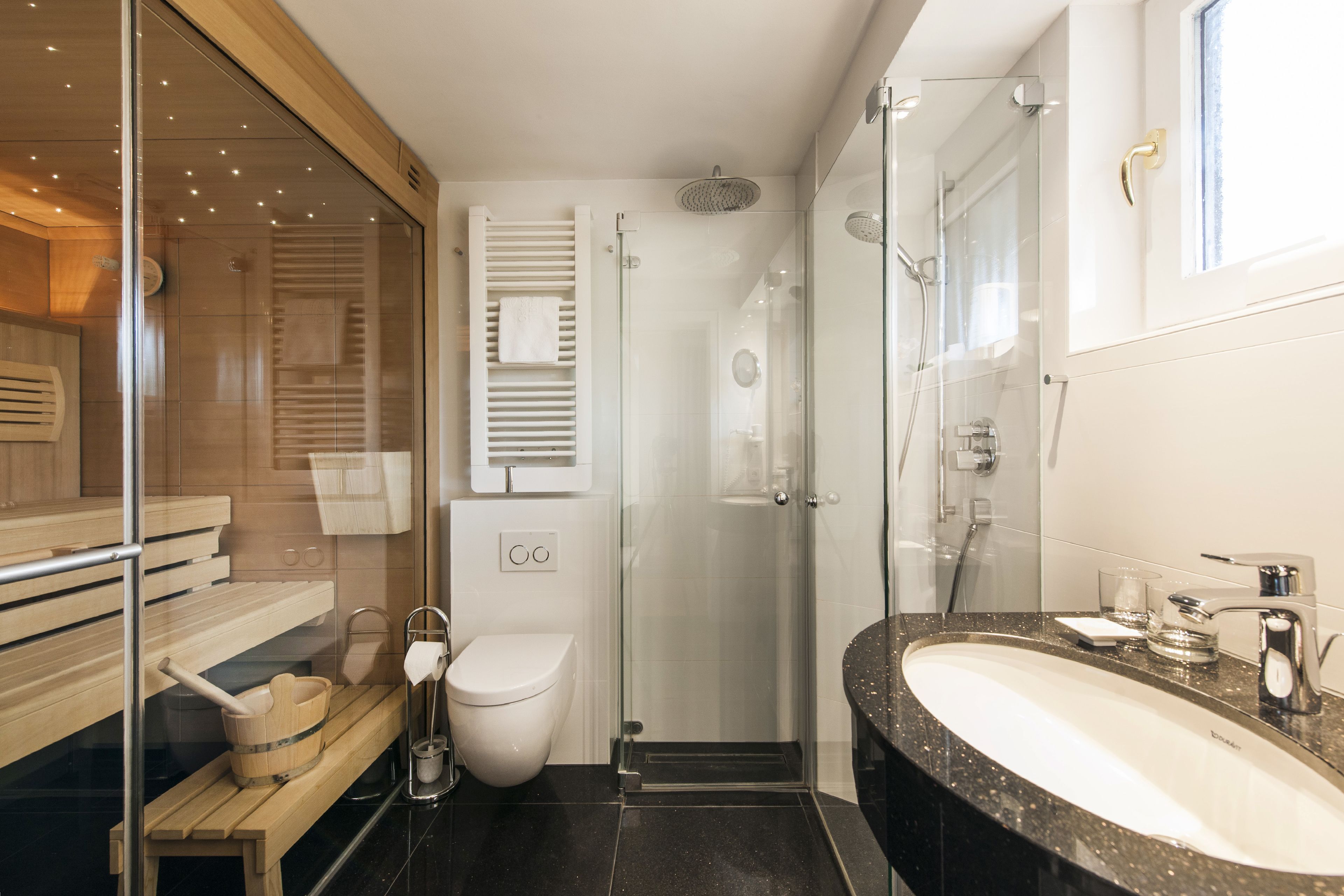 Un baño moderno con cabina de ducha de vidrio, banco de madera, inodoro blanco y lavamanos de granito.