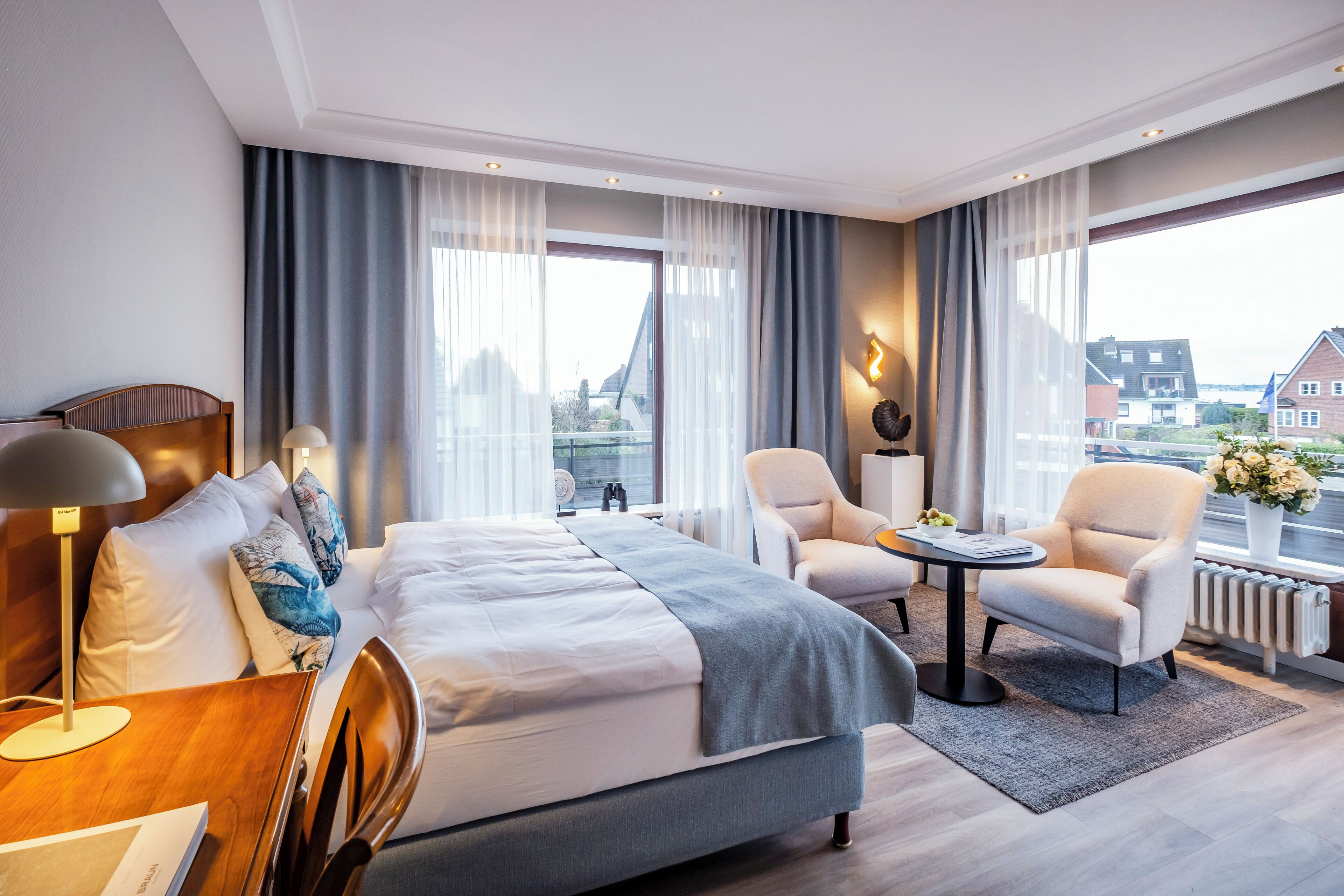 A modern hotel room with a large bed, a desk, two armchairs, and a view of the city.