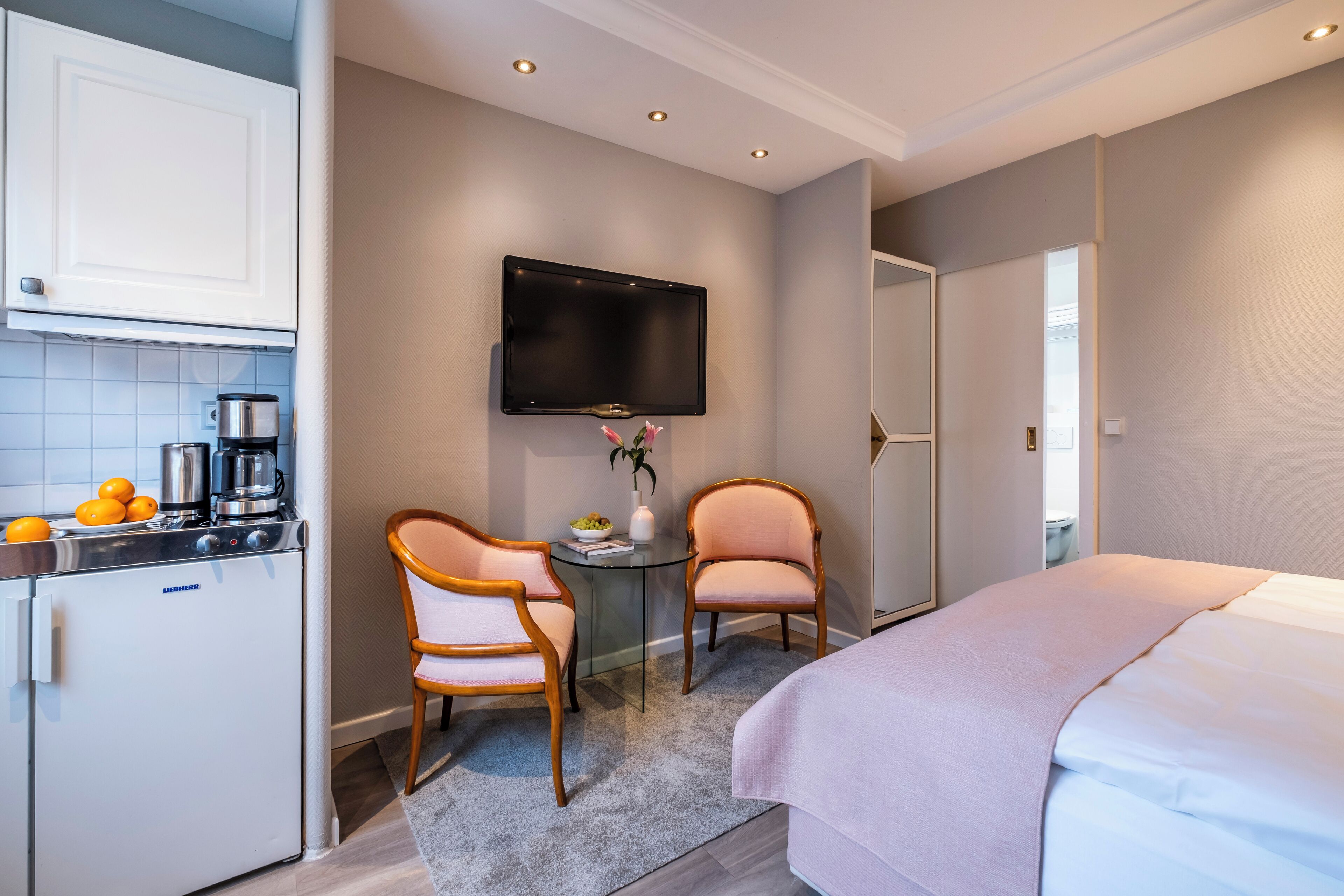 Una habitación de hotel compacta con kitchenette, una cama, televisor y dos sillas.