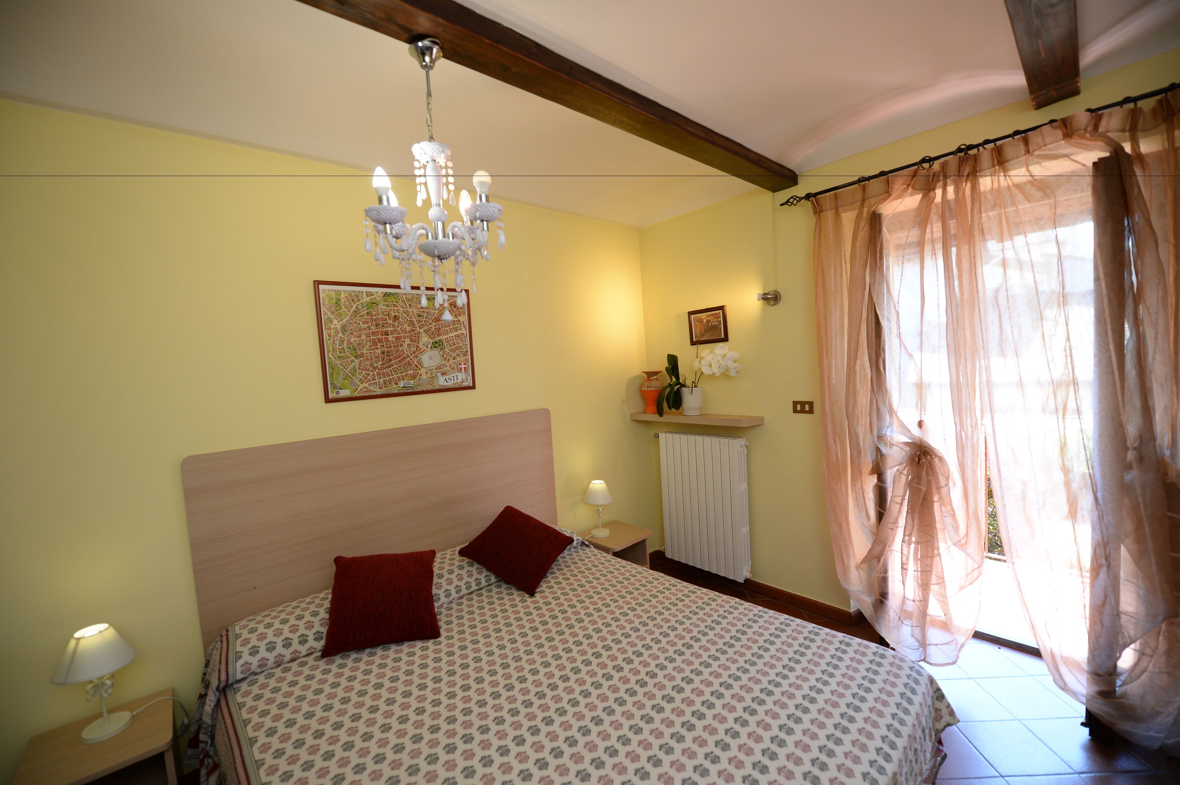 Een slaapkamer met een bed, een kroonluchter, een kaart aan de muur en een raam met dunne gordijnen.
