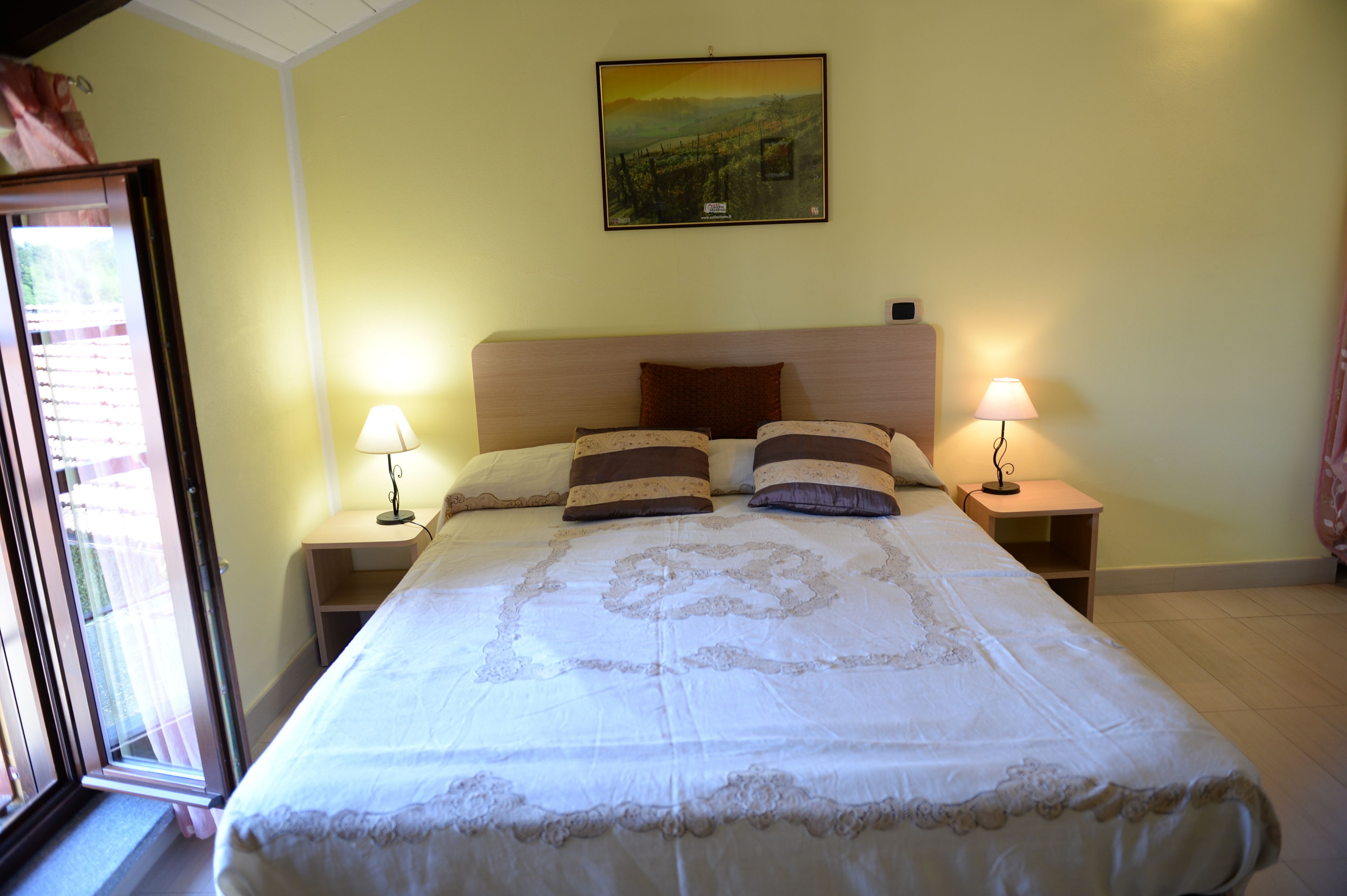 A neatly made bed with a headboard, two bedside tables with lamps, a painting on the wall, and a window with curtains.
