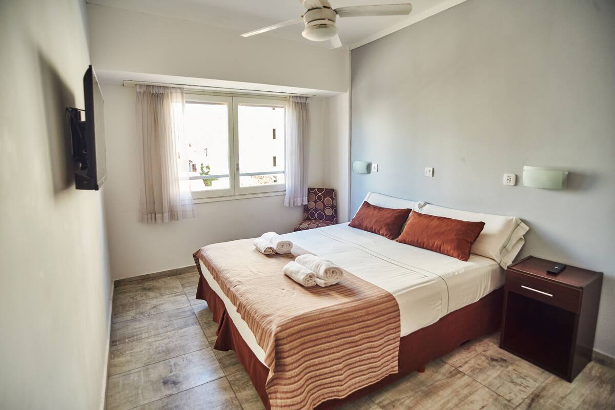 A bedroom with a bed, a nightstand, a ceiling fan, a window with curtains, and a wall-mounted light.