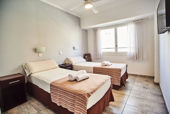 Habitación de hotel con dos camas, un ventilador de techo, una ventana con cortinas y una mesita de noche.