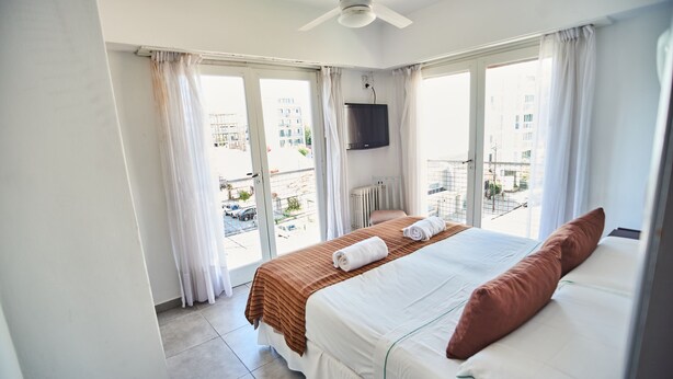 A hotel room with a bed, a ceiling fan, a TV mounted on the wall, and a view of the outside through large windows. - Hotel Pelayo (Mar del Plata)