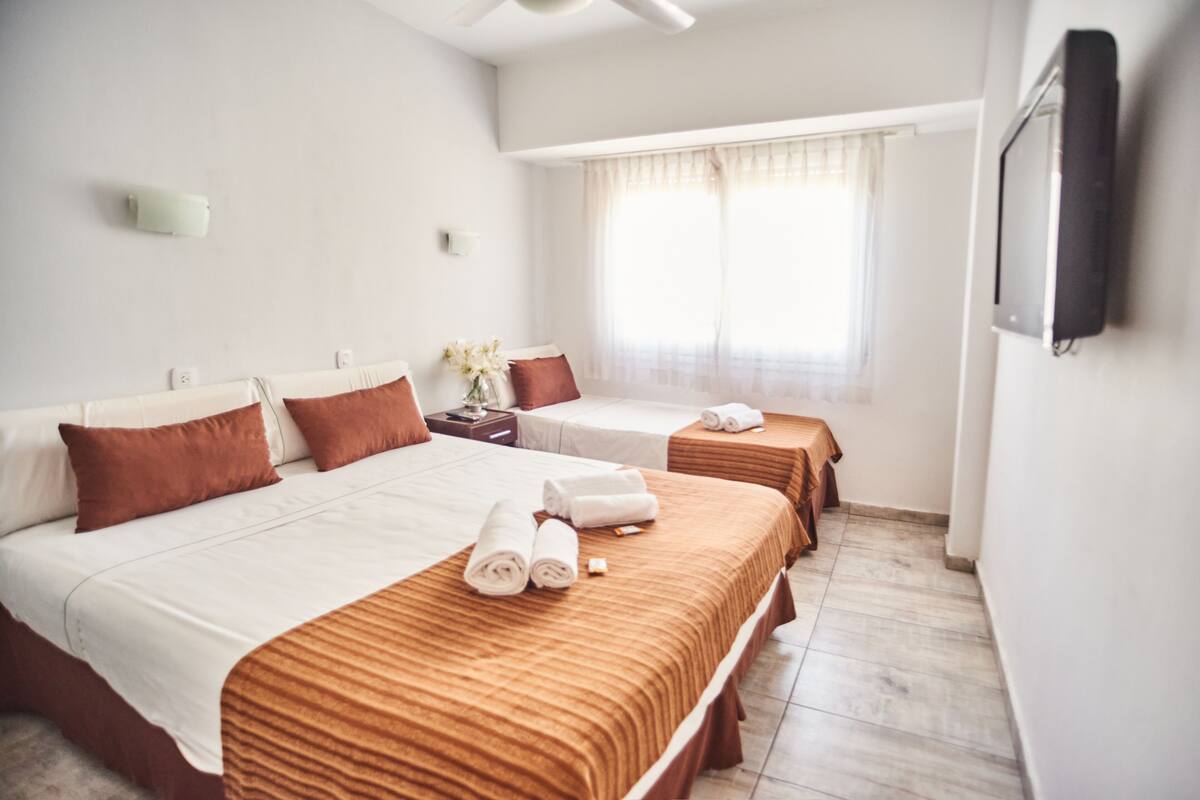 A hotel room with two beds, each with a brown bedspread and white linens. There is a window with sheer curtains, a wall-mounted TV, and a small table with a vase of flowers.