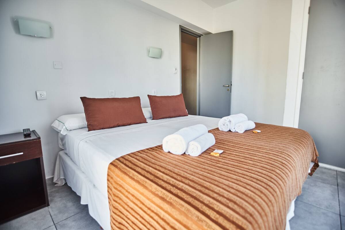 A neatly made bed with white and brown bedding, a wooden nightstand with a remote control, and a pair of rolled towels on the bed.