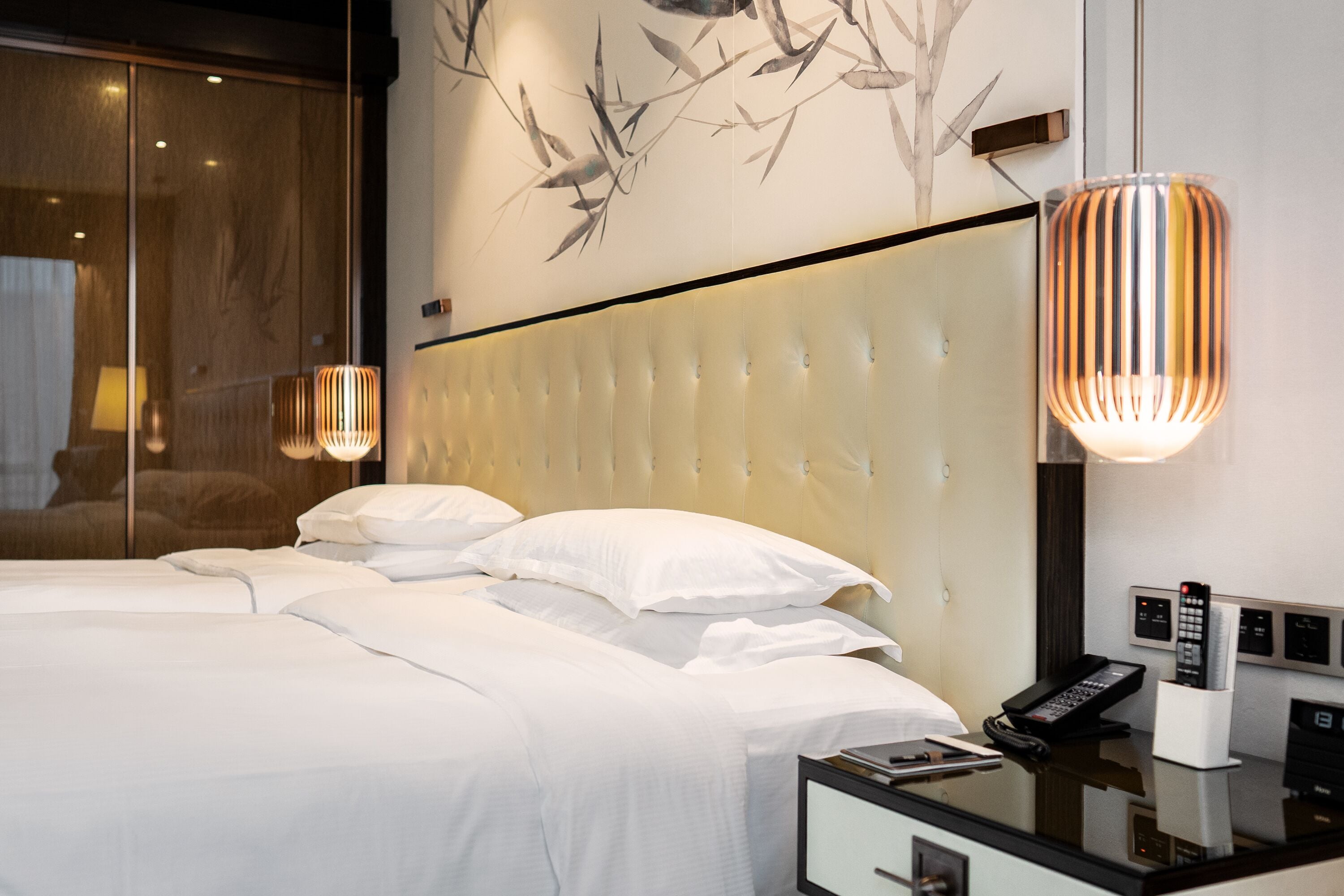 A modern hotel room with a large bed, a bedside table with a phone and remote controls, and a wall with a bamboo leaf mural.