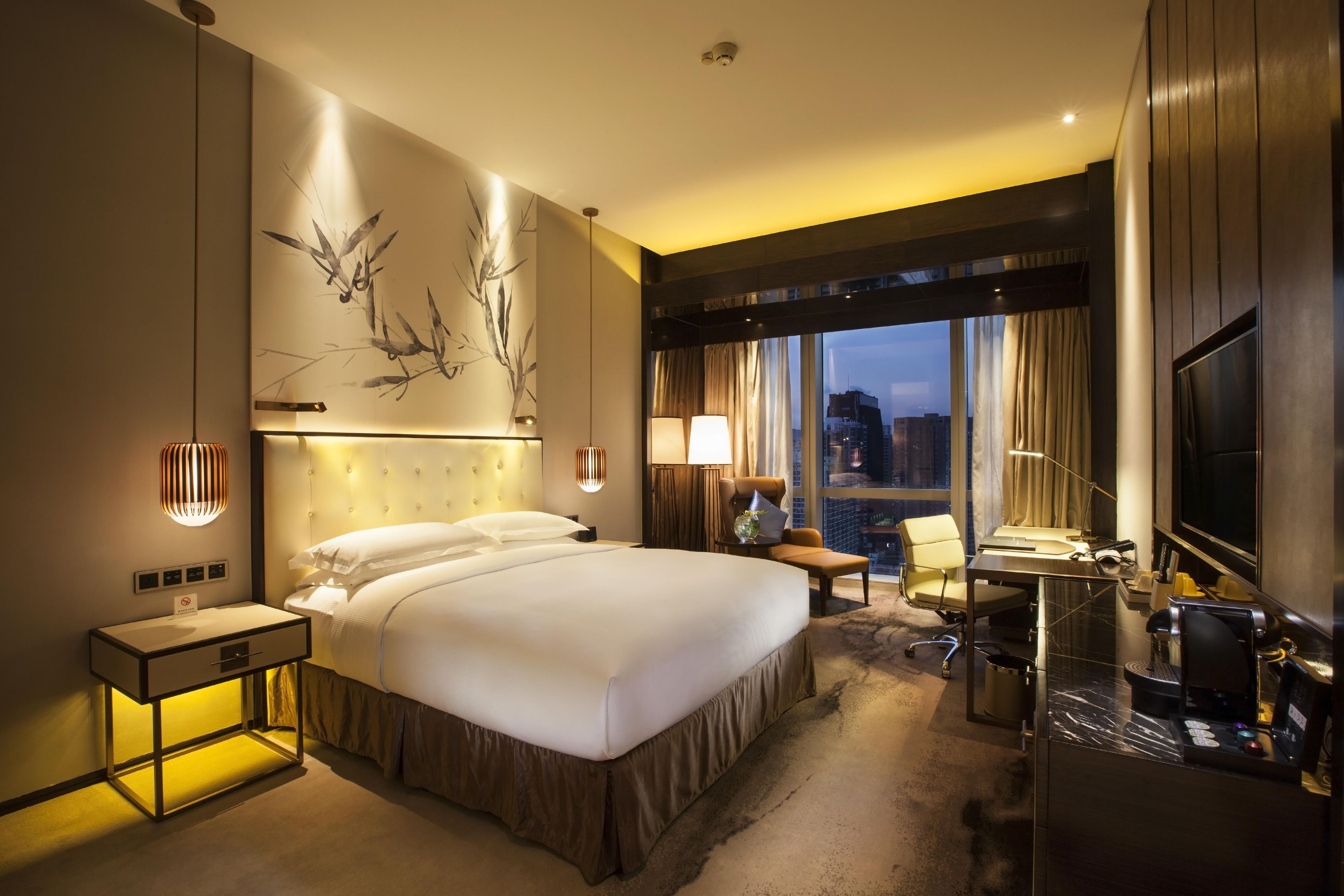 A hotel room with a large bed, bedside tables, a desk, a chair, a TV, and a view of the cityscape.