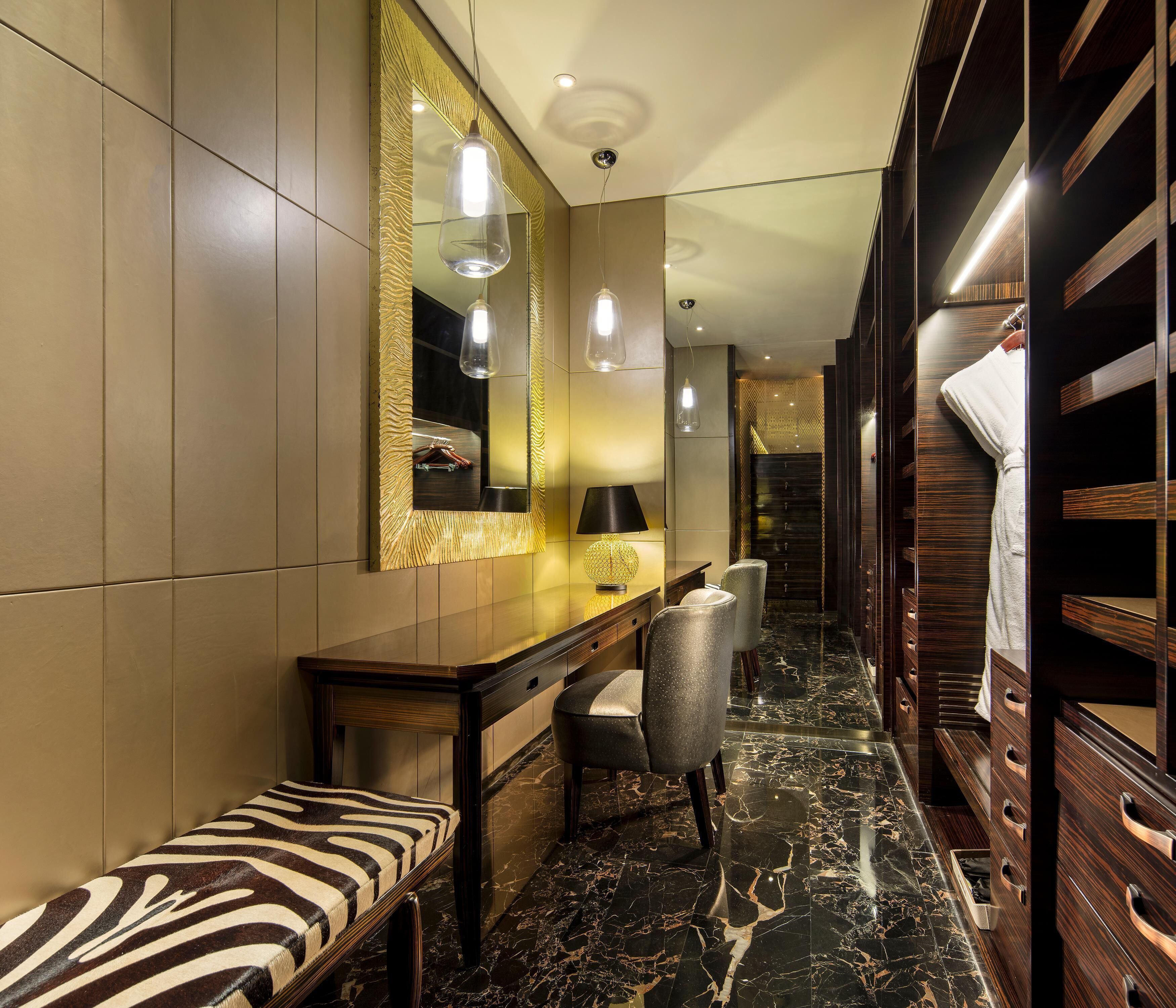 Ein Hotelzimmer mit Marmorboden, einer Bank mit Zeprazierkissen, einem Schreibtisch mit Lampe, einem Spiegel mit goldener Rahmung und einem Holzschrank.