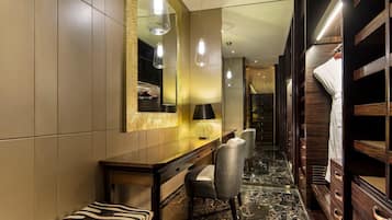 Ein Hotelzimmer mit Marmorboden, einer Bank mit Zeprazierkissen, einem Schreibtisch mit Lampe, einem Spiegel mit goldener Rahmung und einem Holzschrank.