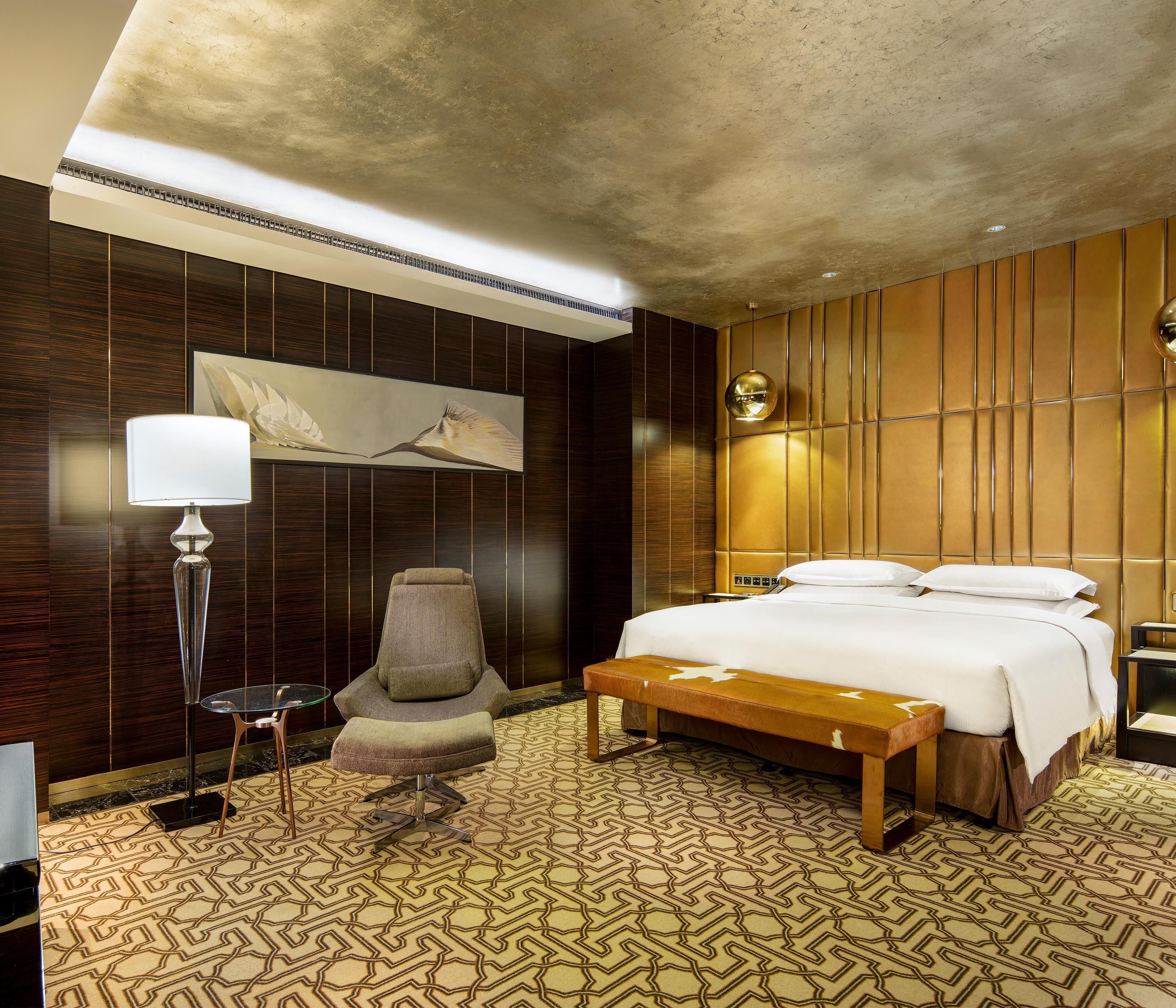 A hotel room with a bed, a chair, a table, a lamp, and a wall-mounted artwork.