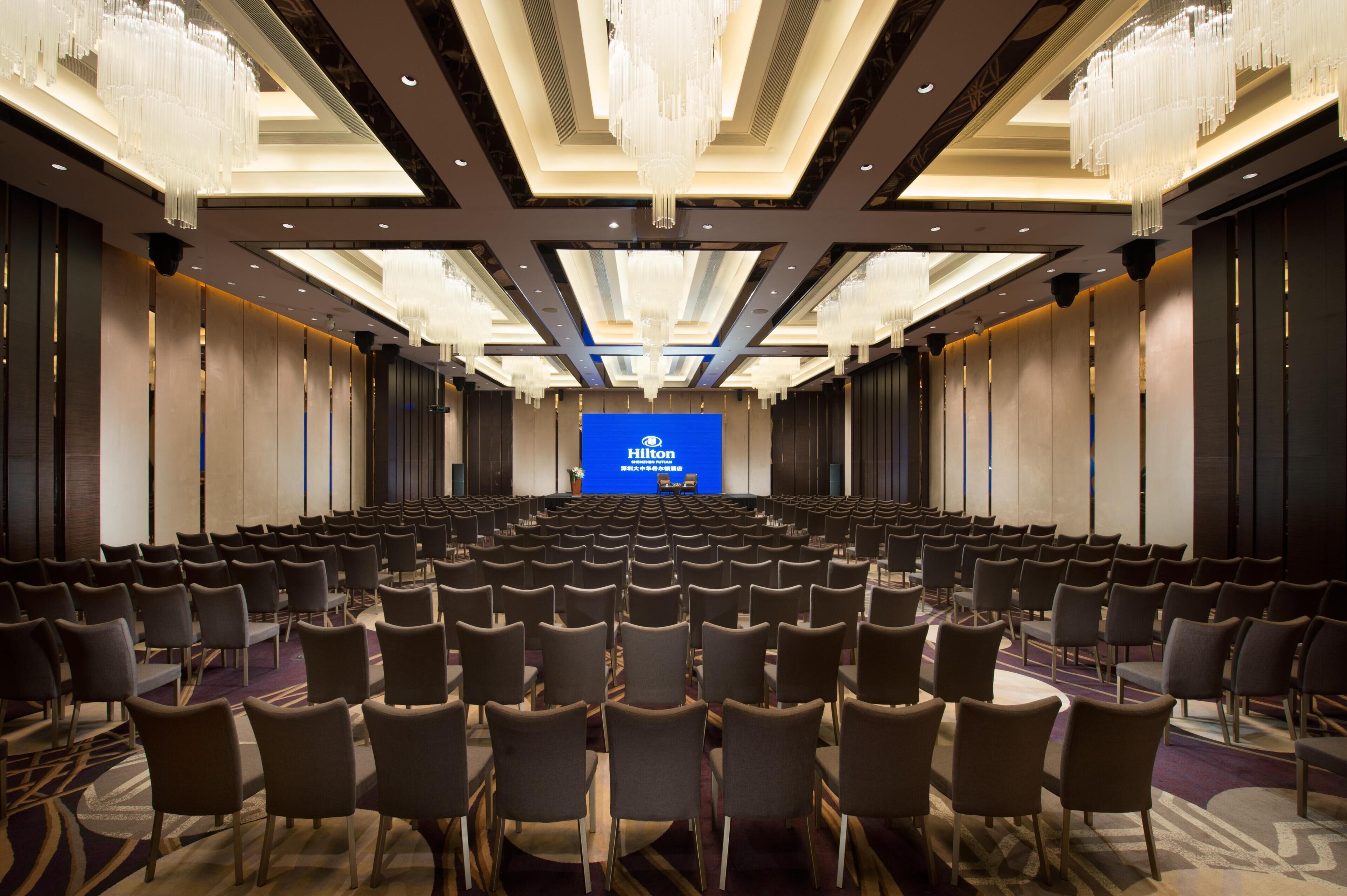 Uma sala de conferências com fileiras de cadeiras, uma tela grande exibindo o logotipo da Hilton e um lustre moderno.