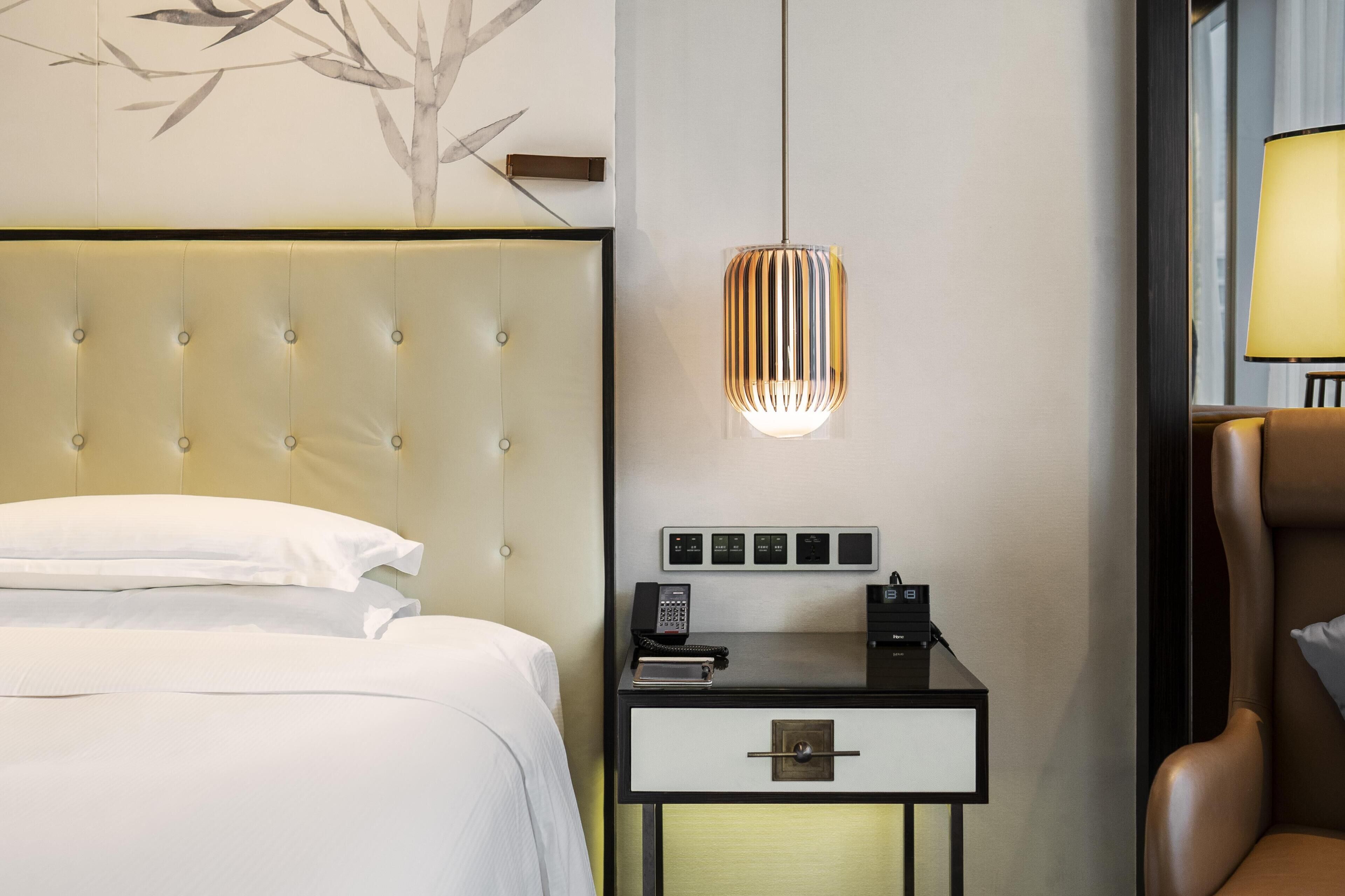 A hotel room with a tufted headboard, a bedside table with a phone and a lamp, and a chair with a cushion.