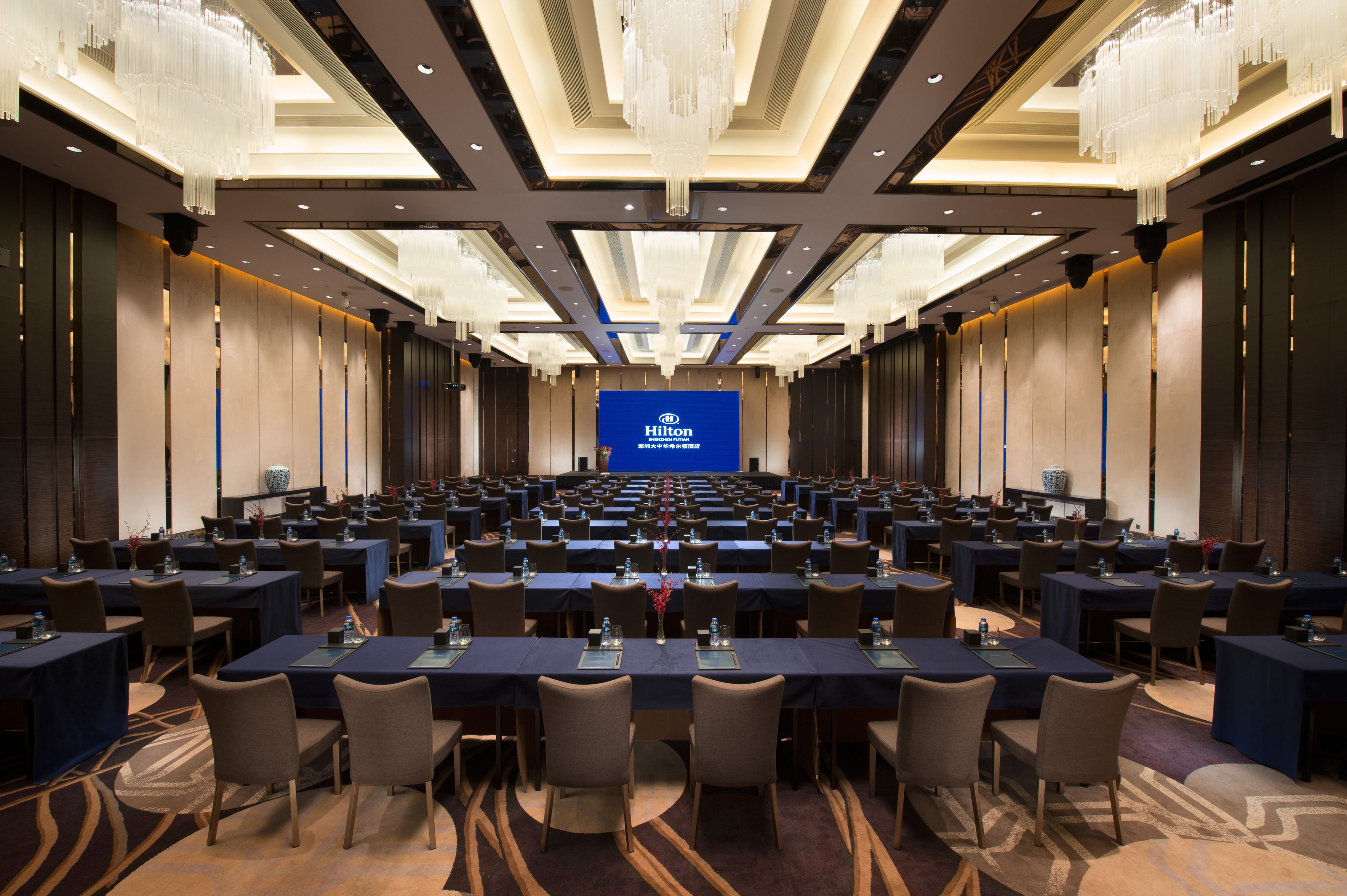 Un amplio salón de conferencias con filas de mesas y sillas, una pantalla azul con el logo de Hilton y candelabros modernos.