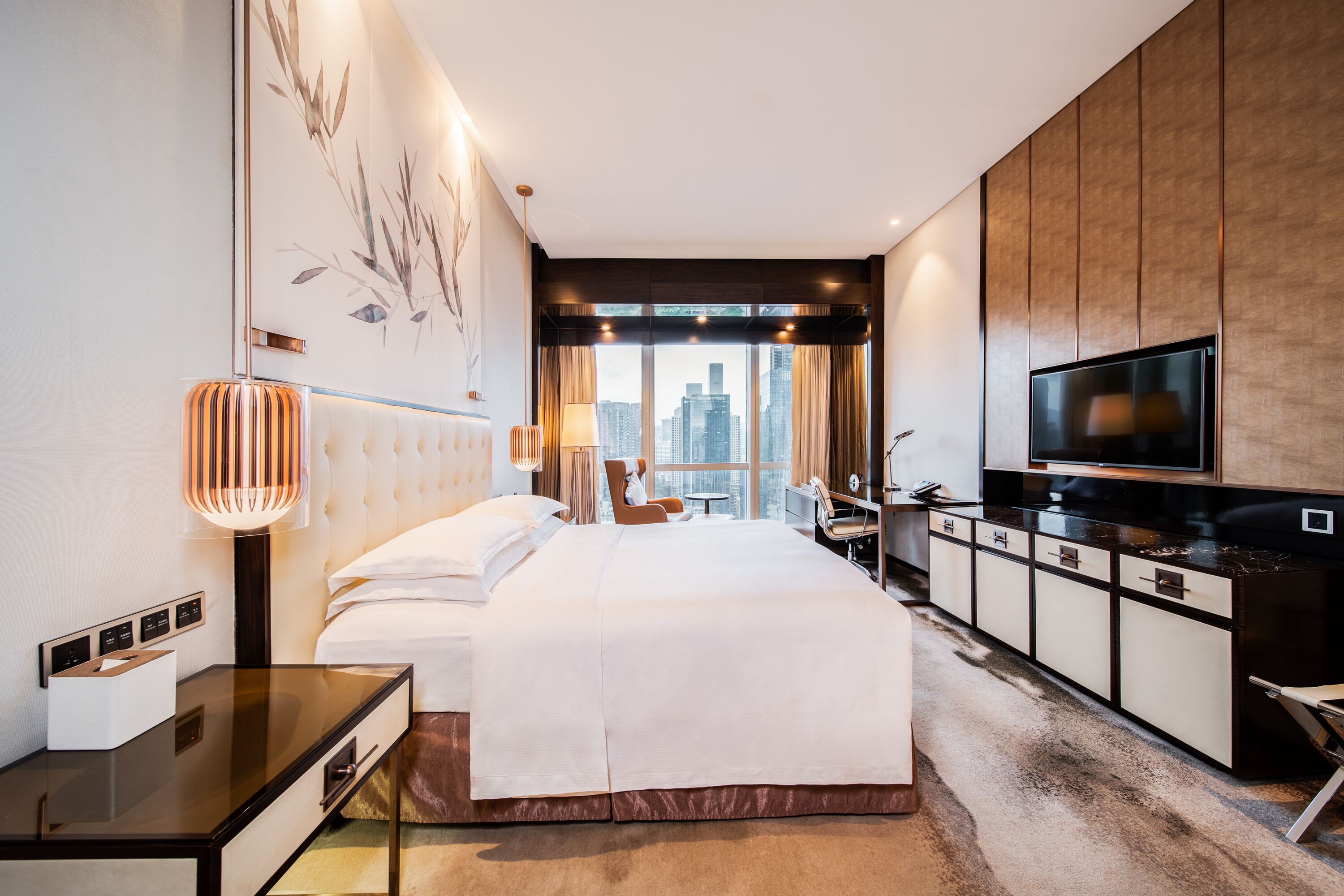 A modern hotel room with a large bed, a desk, a television, and a view of the cityscape.