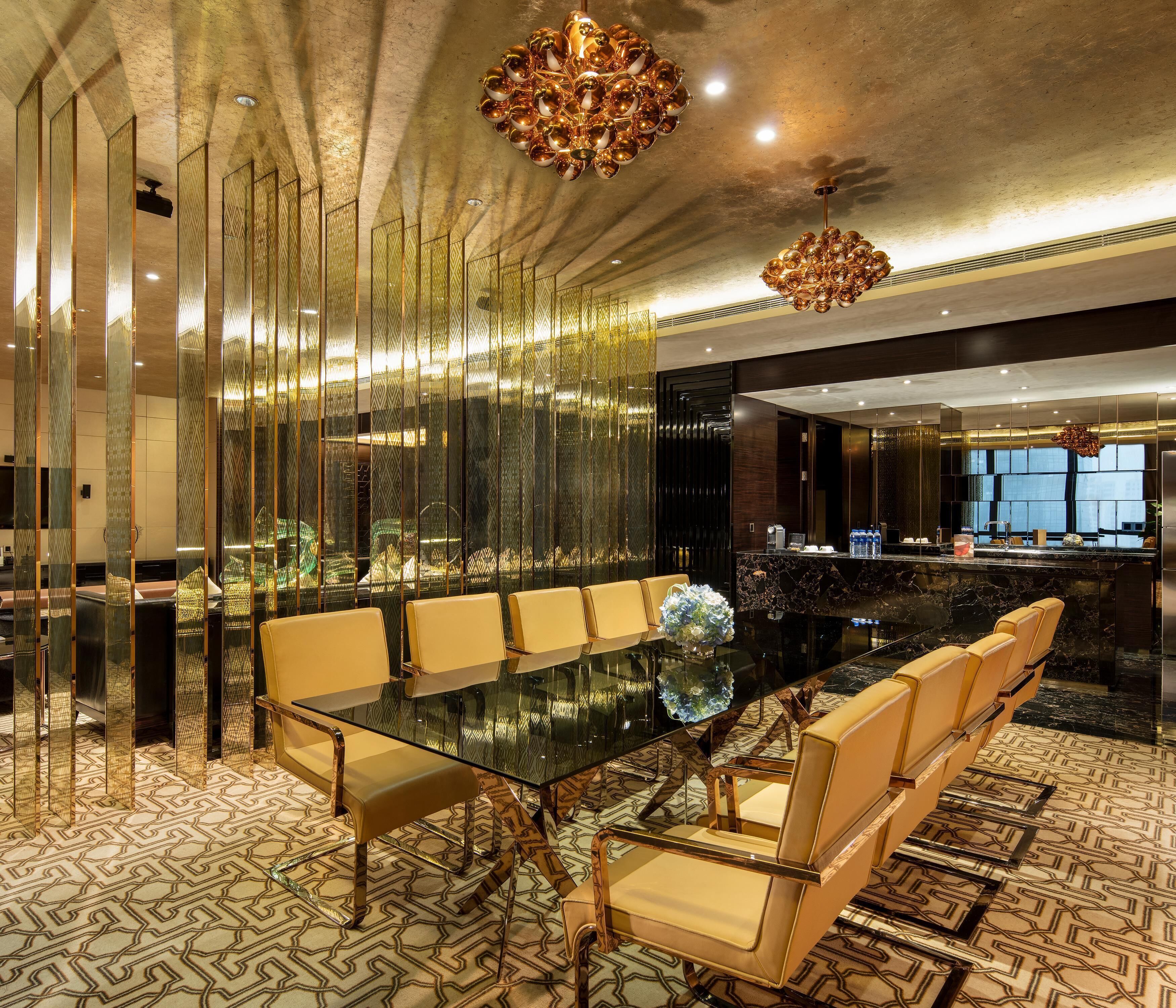 A modern hotel lobby with a glass-top dining table, leather chairs, a decorative chandelier, and a reception desk.
