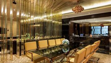 A modern hotel lobby with a glass-top dining table, leather chairs, a decorative chandelier, and a reception desk.