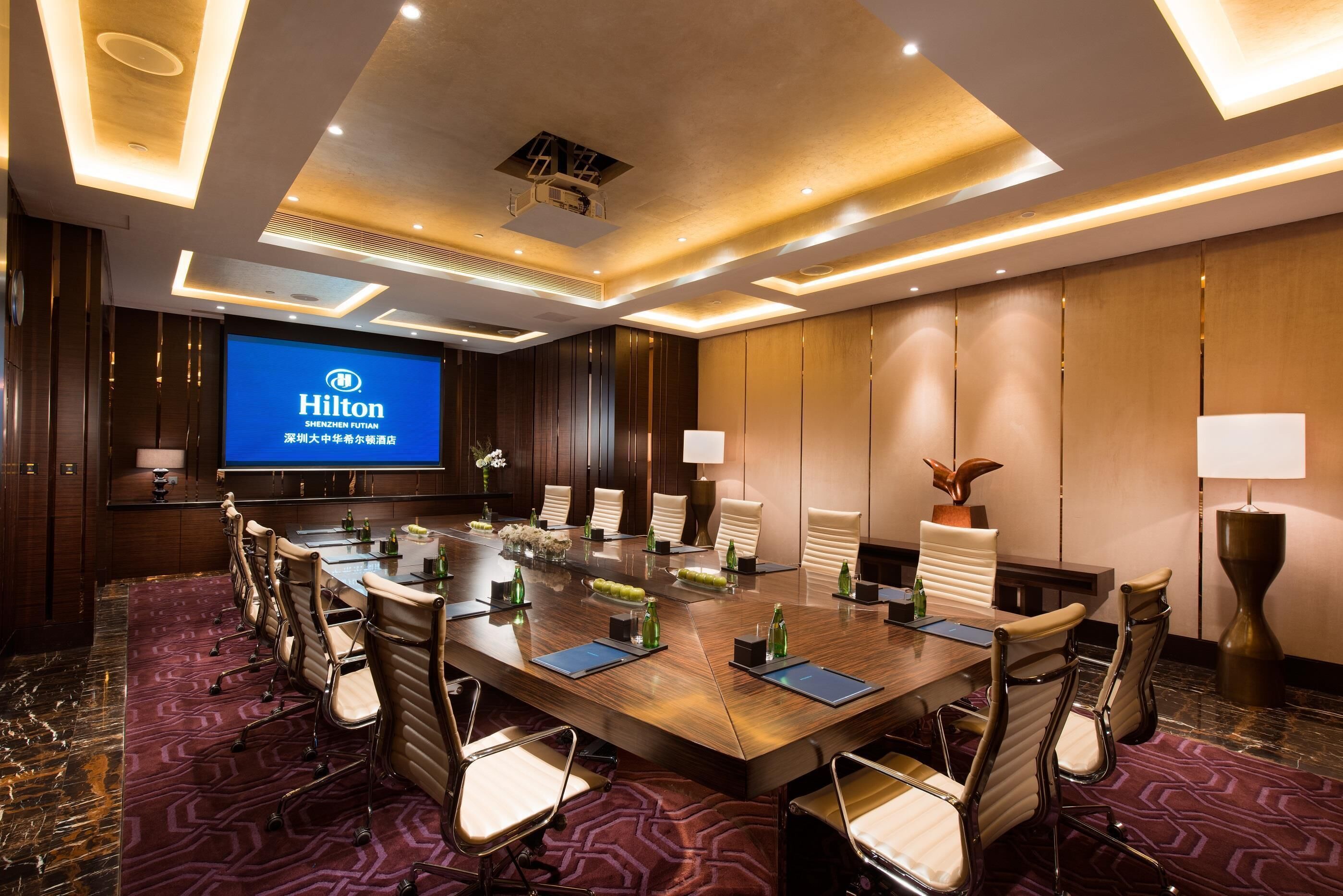 A conference room with a long table, chairs, a large screen displaying the Hilton logo, and a modern ceiling design.