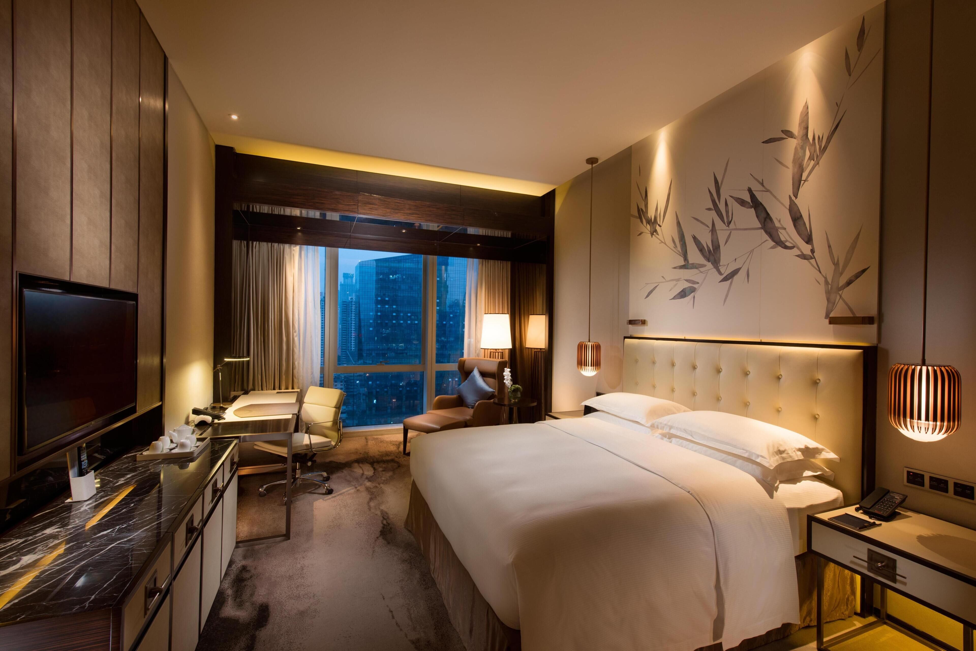 Ein Hotelzimmer mit einem großen Bett, einem Schreibtisch mit Fernseher, einem Sessel, einem Fenster mit Stadtblick und einer Wand mit Bambusmuster.