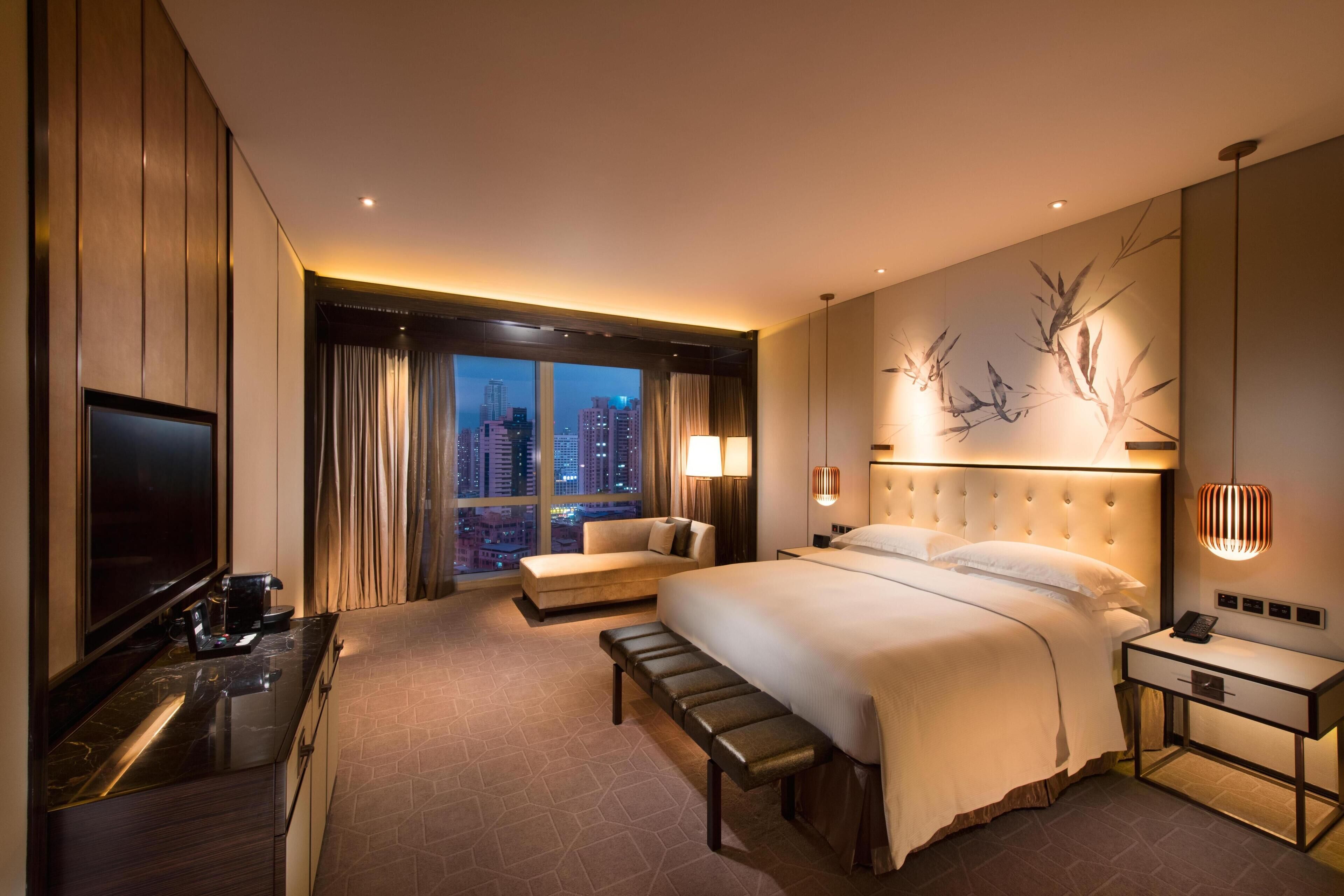 A hotel room with a large bed, a sofa, a desk with a TV, and a city view.