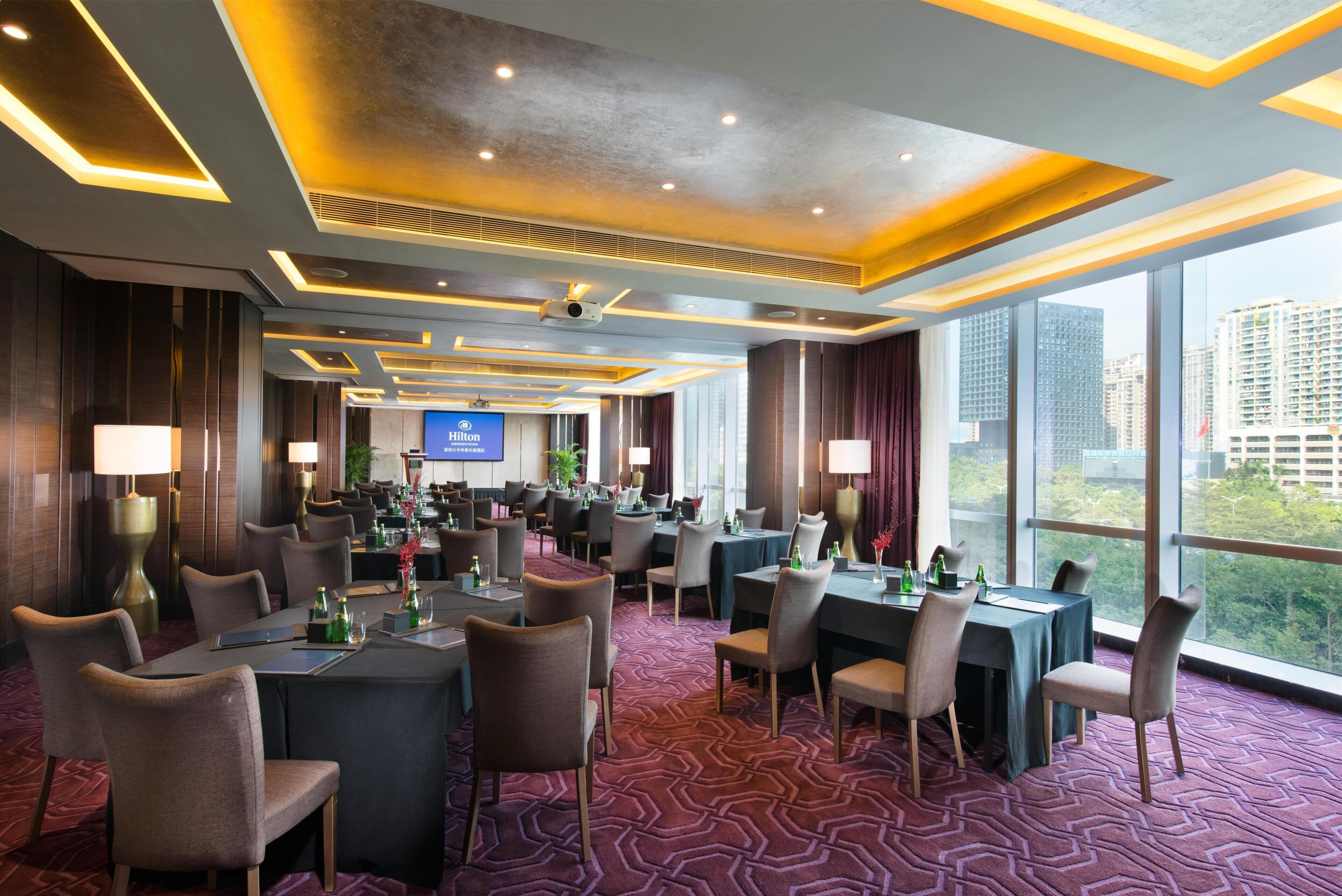 A hotel conference room with multiple tables and chairs, a large screen, and a view of the cityscape through large windows.