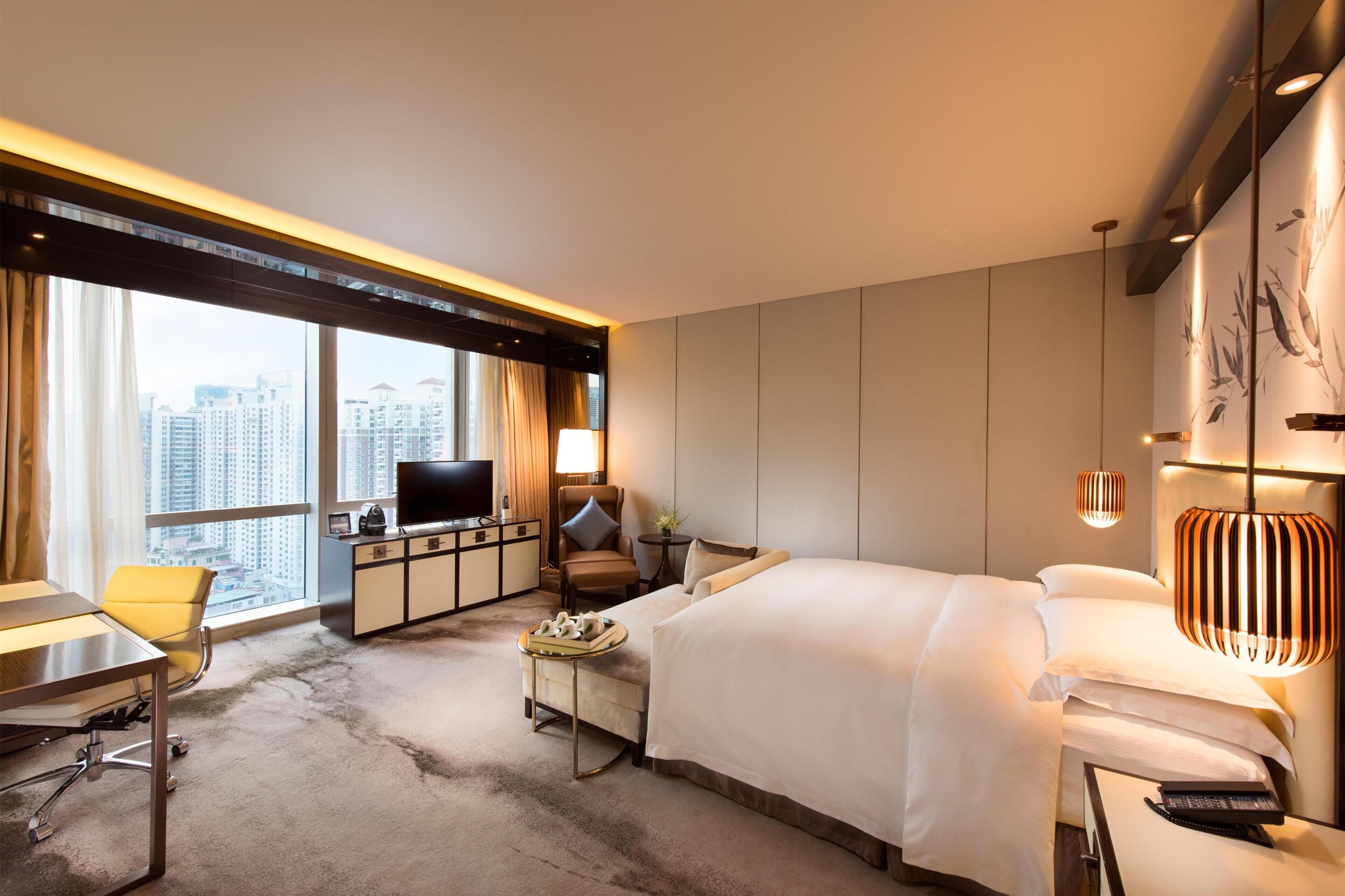 A hotel room with a large bed, a desk with a chair, a television, and a view of the cityscape through large windows.