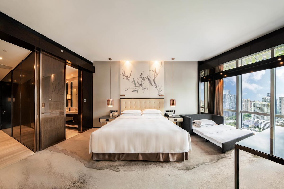 A modern bedroom with a large bed, a sofa bed, a walk-in closet, and a city view.