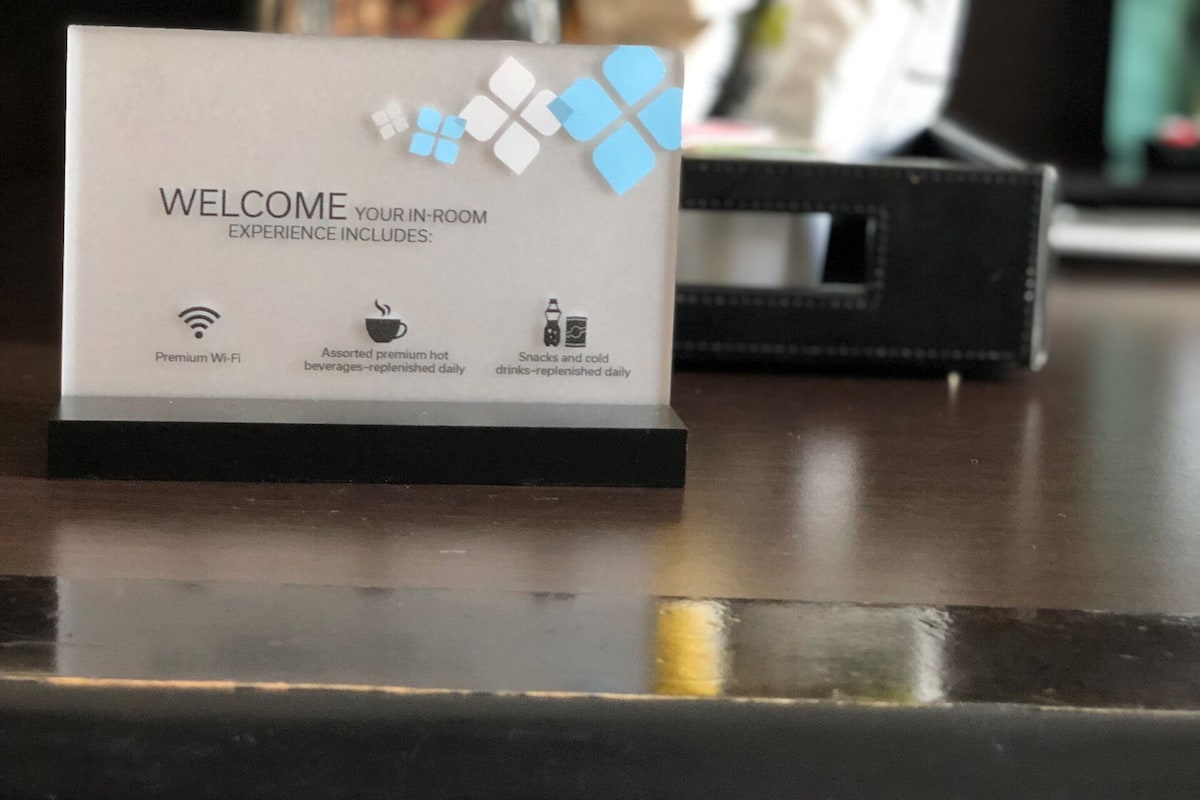 Señal de bienvenida que detalla los servicios: Wi-Fi de alta velocidad, variedad de bebidas calientes premium, y snacks y bebidas frías reabastecidos diariamente.