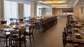 Une salle à manger d'hôtel avec de nombreuses tables et chaises, un bar en arrière-plan et de grandes fenêtres avec des rideaux.
