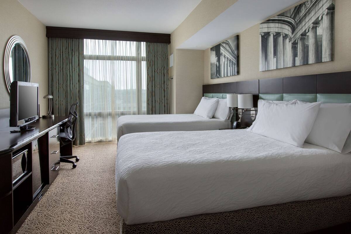 Habitación de hotel con dos camas, un escritorio con computadora, una silla, un televisor y ventanas grandes con cortinas.