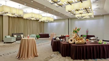 Un salón de conferencias con mesas redondas cubiertas con manteles marrones, un bufé con diversas opciones de comida y lámparas modernas.