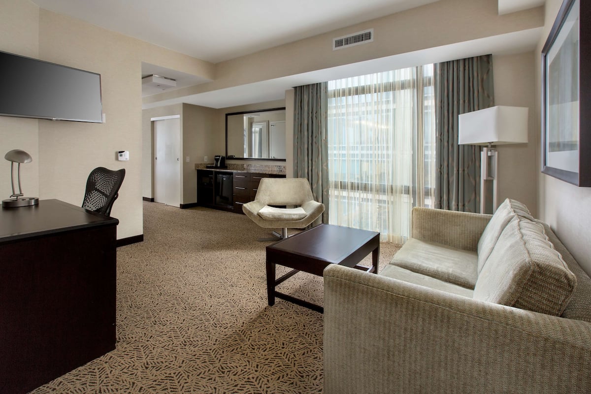 Habitación de hotel con televisión de pantalla plana, escritorio, sofá, sillón y comedor con mostrador de cocina.
