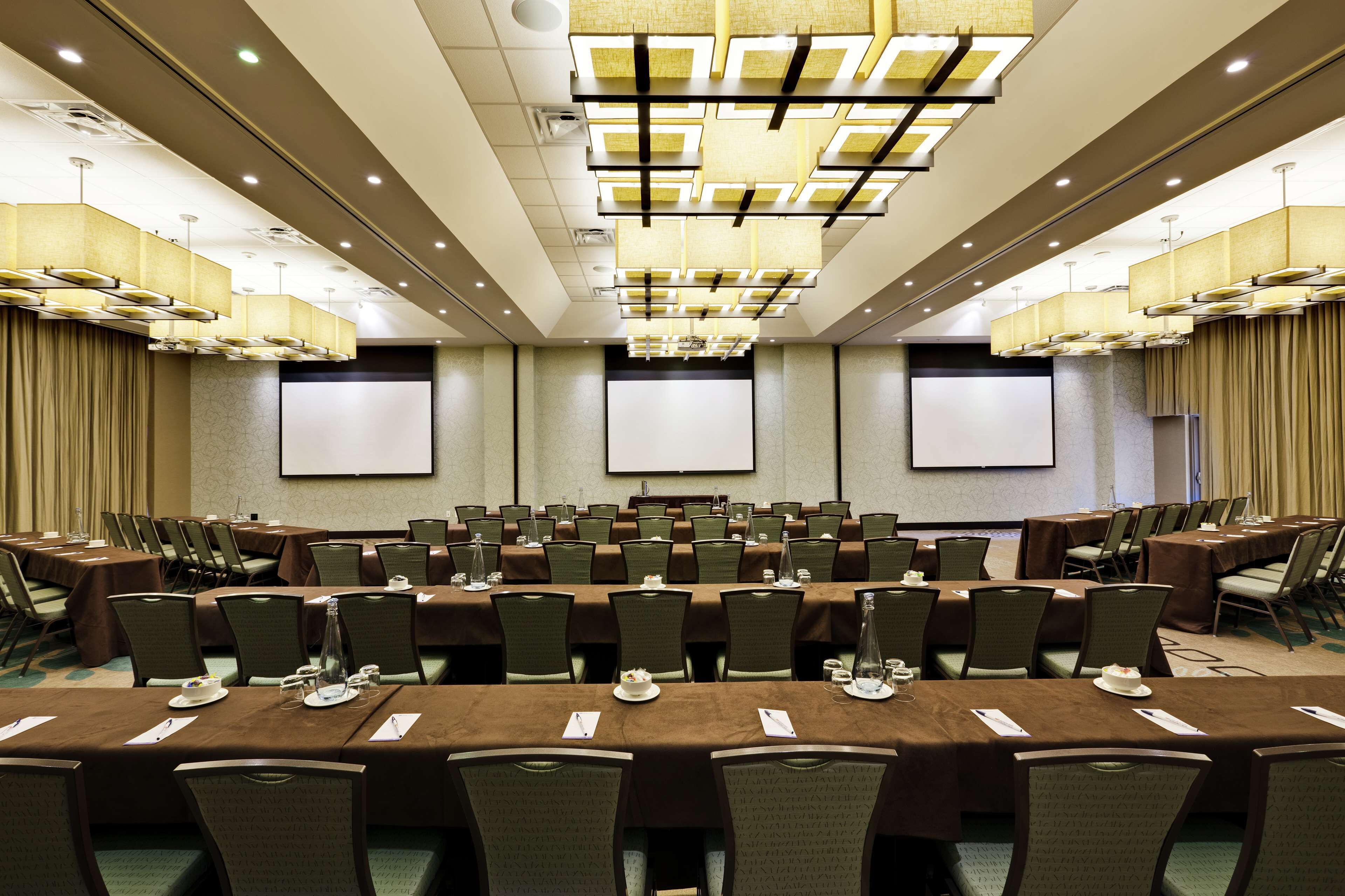 Un salón de conferencias con varias mesas y sillas, equipado con pantallas de presentación e iluminación colgante.