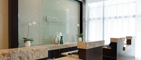 Un moderno mostrador de recepción de hotel con un muro de cristal que muestra el logo de Hilton Garden Inn, una encimera de mármol y plantas en macetas.
