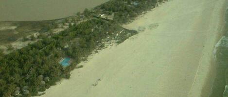 An aerial view of a beach with a long stretch of sand, a few scattered buildings, and a body of water to the left.