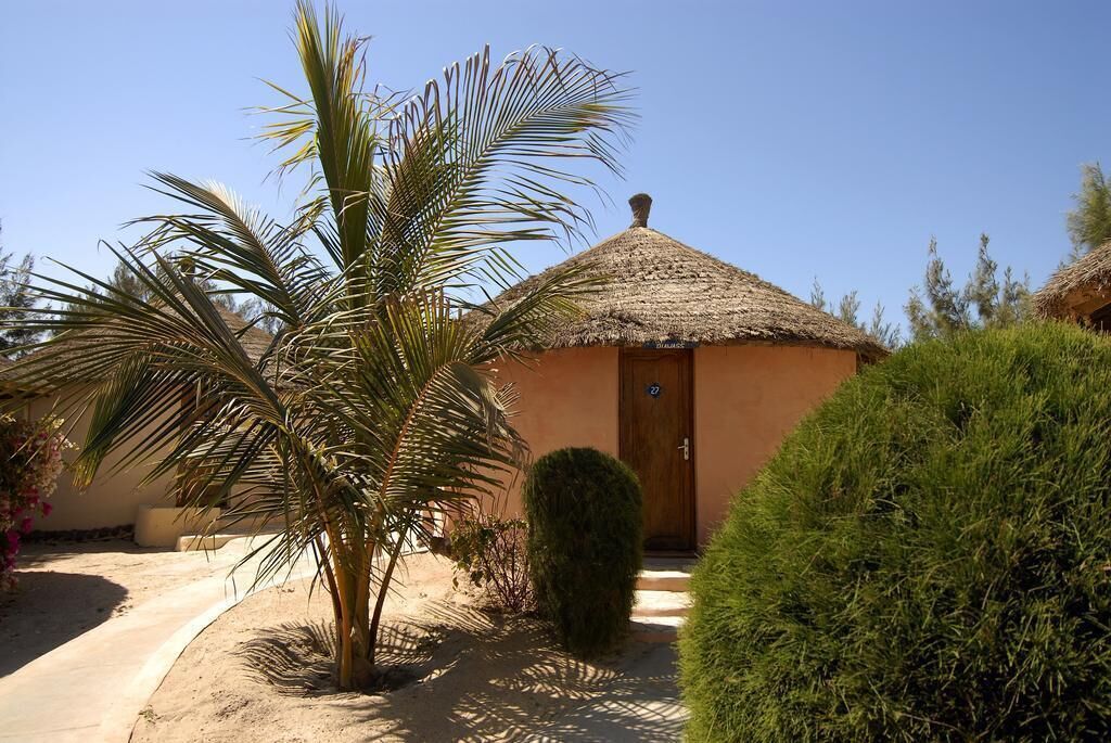 Un edificio pequeño de una sola planta con techo de paja, una puerta con número y una palmera en primer plano.