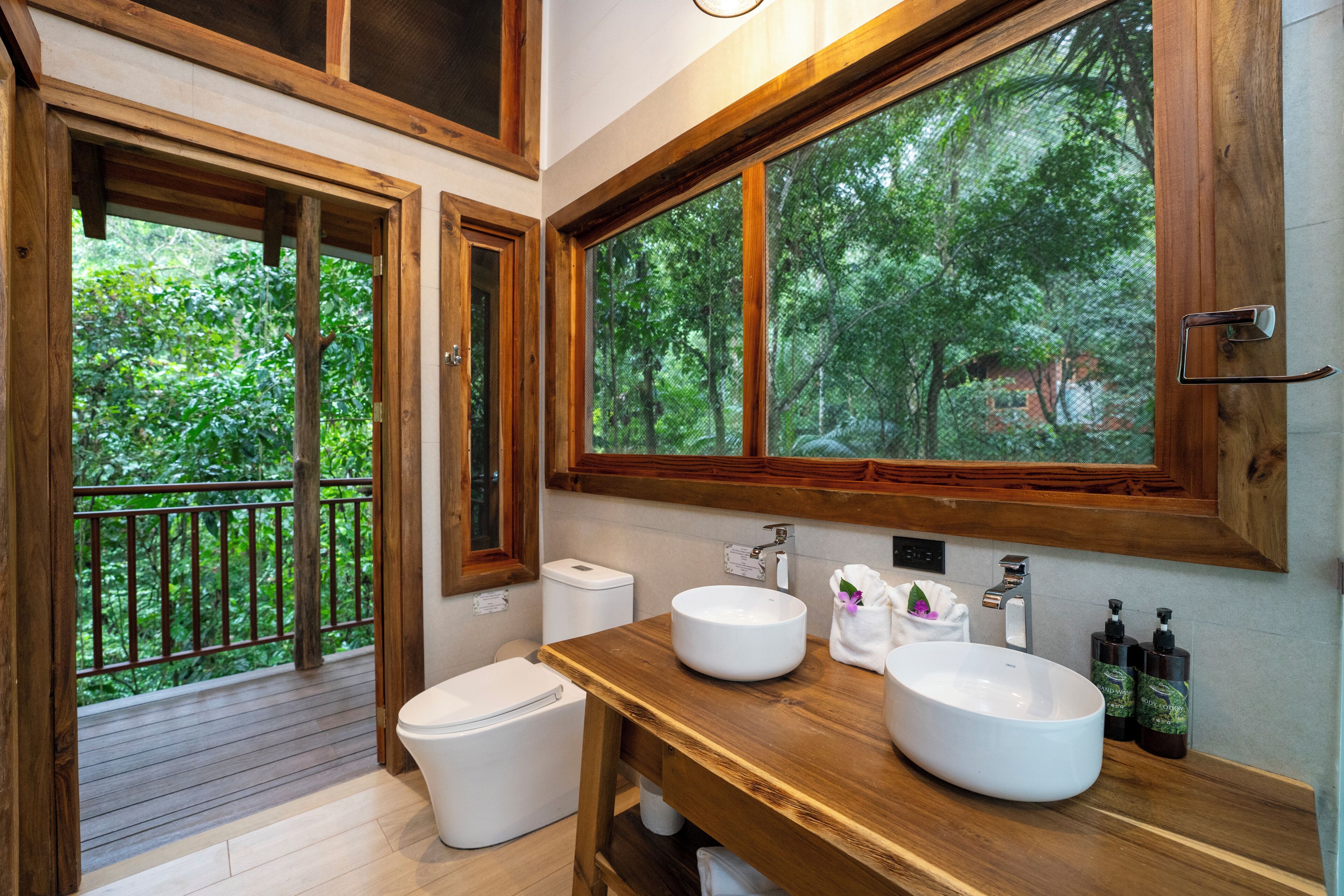 Un baño con tocador de madera, dos lavamanos, un inodoro y un ventanal con vistas a árboles.