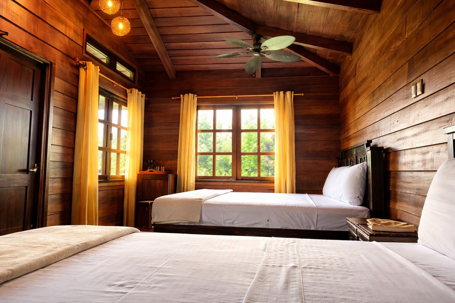 Une chambre dans une cabane en bois, avec deux lits, un ventilateur de plafond et de grandes fenêtres avec des rideaux.