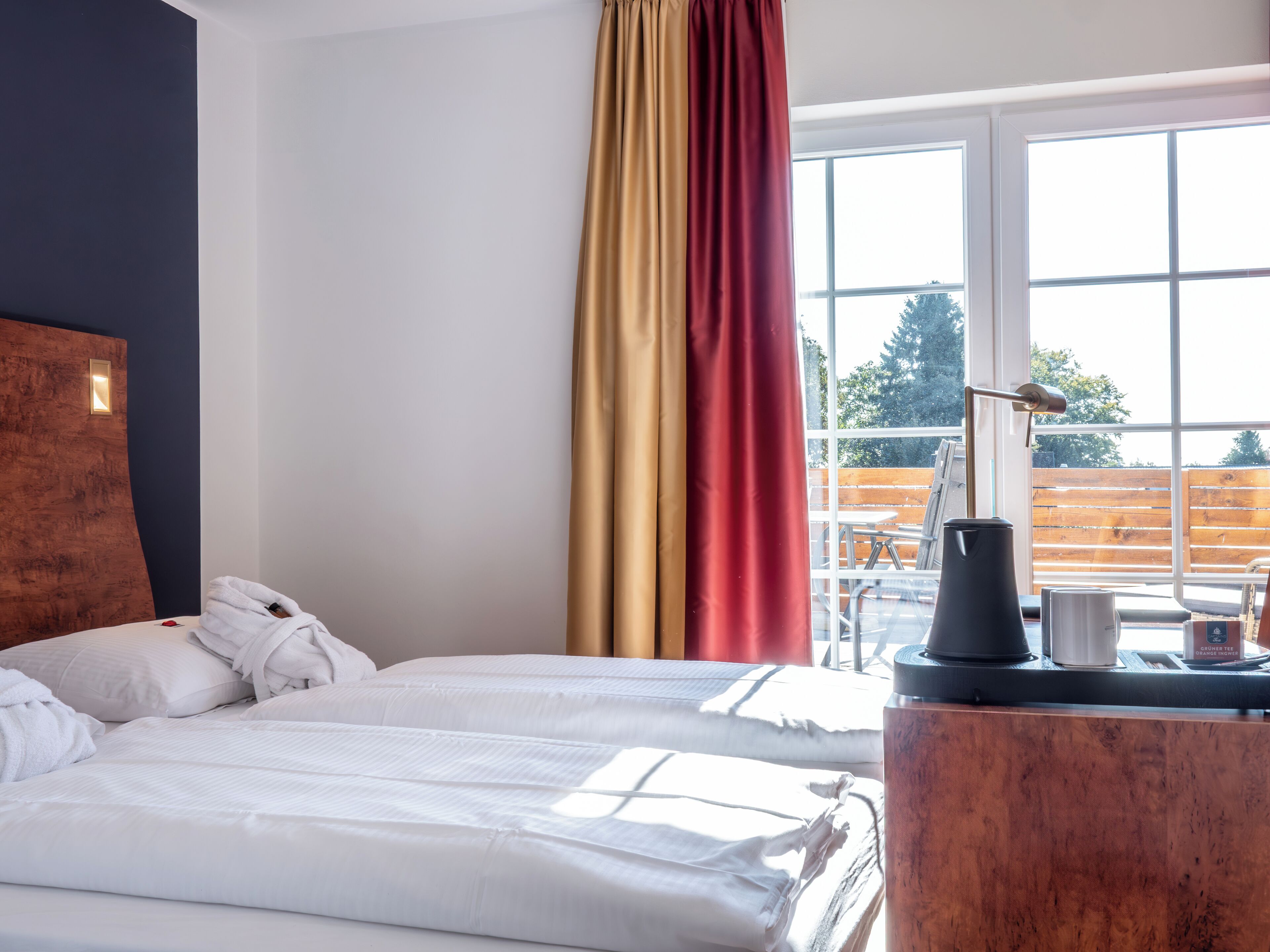 Habitación de hotel con cama, cabecera de madera, ventana con cortinas y mesa con cafetera.