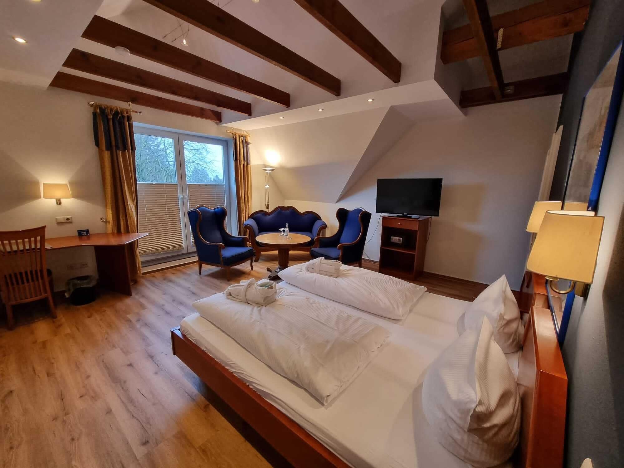 Habitación de hotel con una cama grande, un escritorio con una silla, una pequeña mesa con dos sillas, un televisor y una ventana con cortinas.